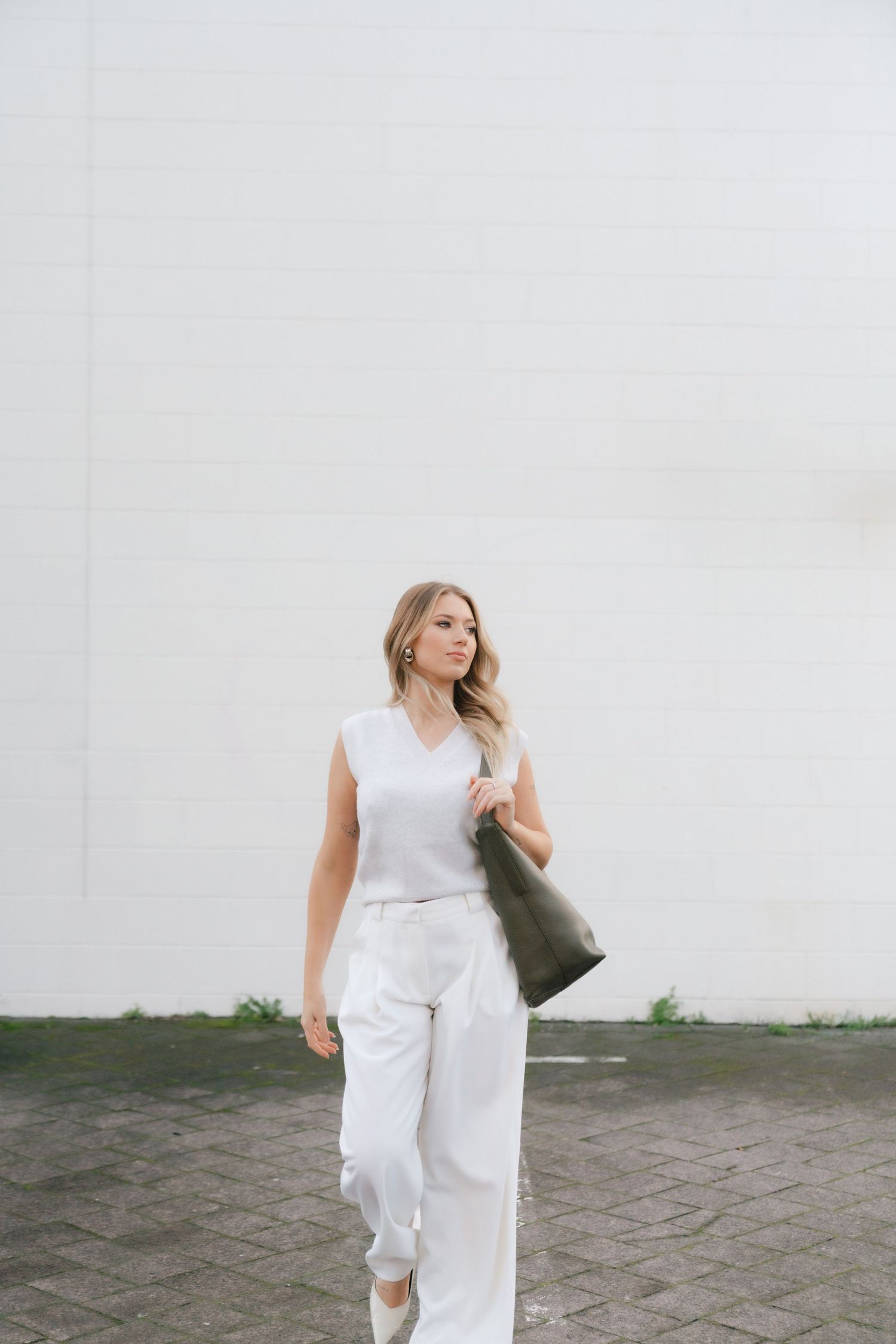 Woman in white outfit and wavy blonde hair walking with green bag against neutral wall background.