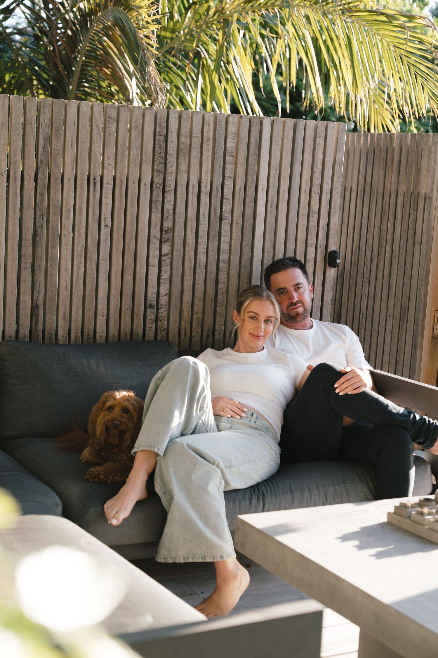A couple and their dog relax together on outdoor patio furniture with palm trees visible behind a wooden fence.