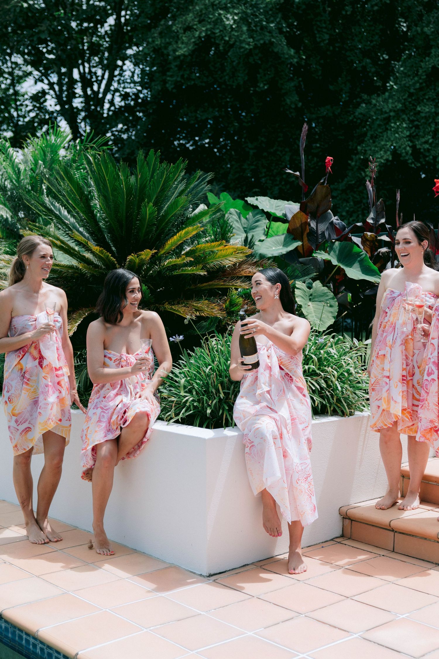 Four attendants in floral pink dresses gather by a poolside patio surrounded by lush tropical plants.