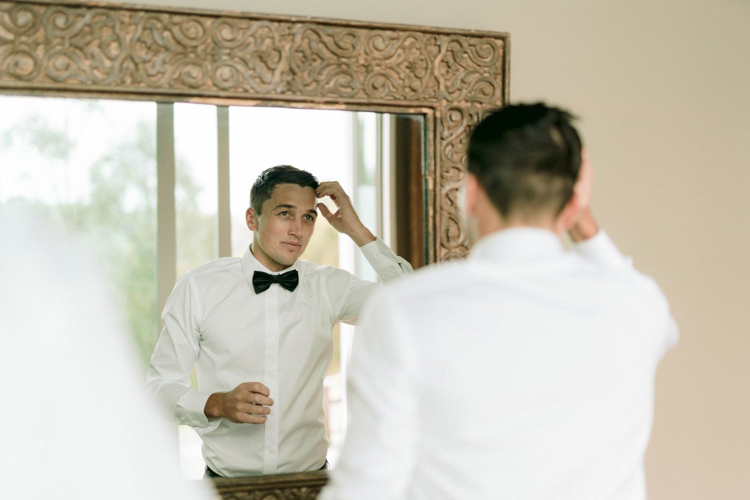 Individual adjusting formal attire while looking in ornate framed mirror.