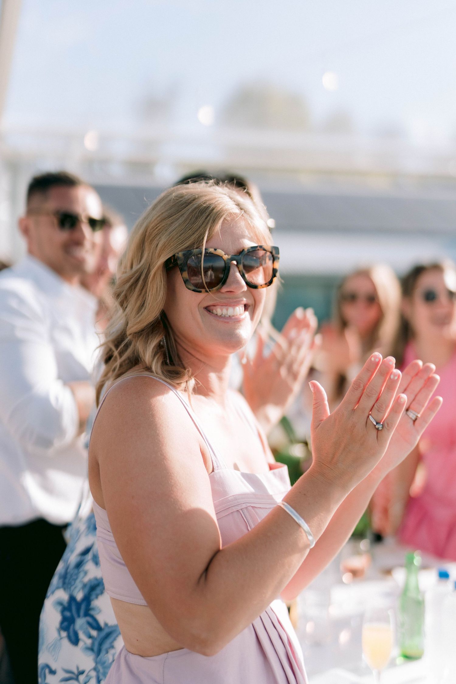 A guest in a light pink dress and sunglasses claps and smiles during an outdoor event.