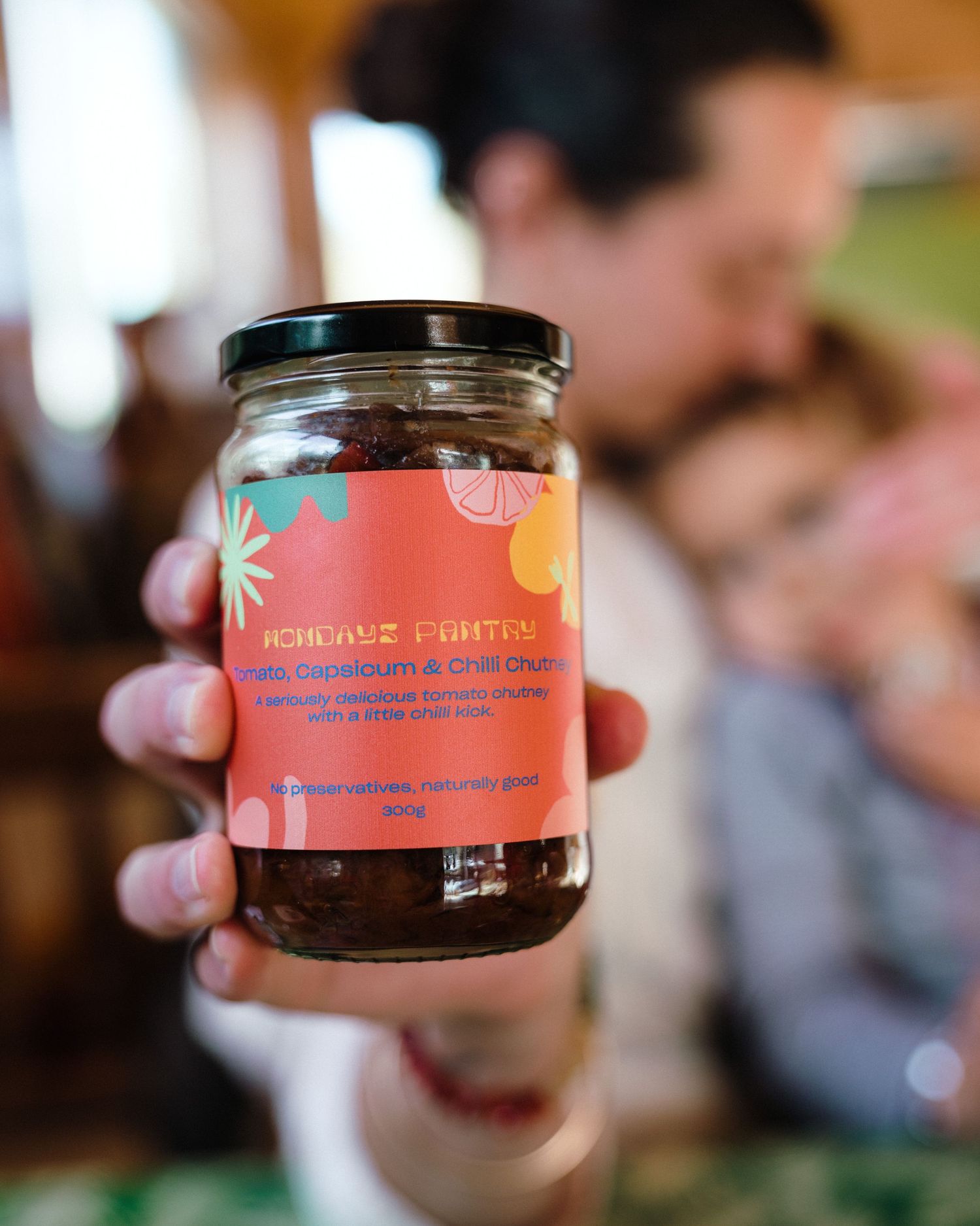 Hand holding a jar of artisanal fruit spread with colorful tropical-themed label design against blurred background.