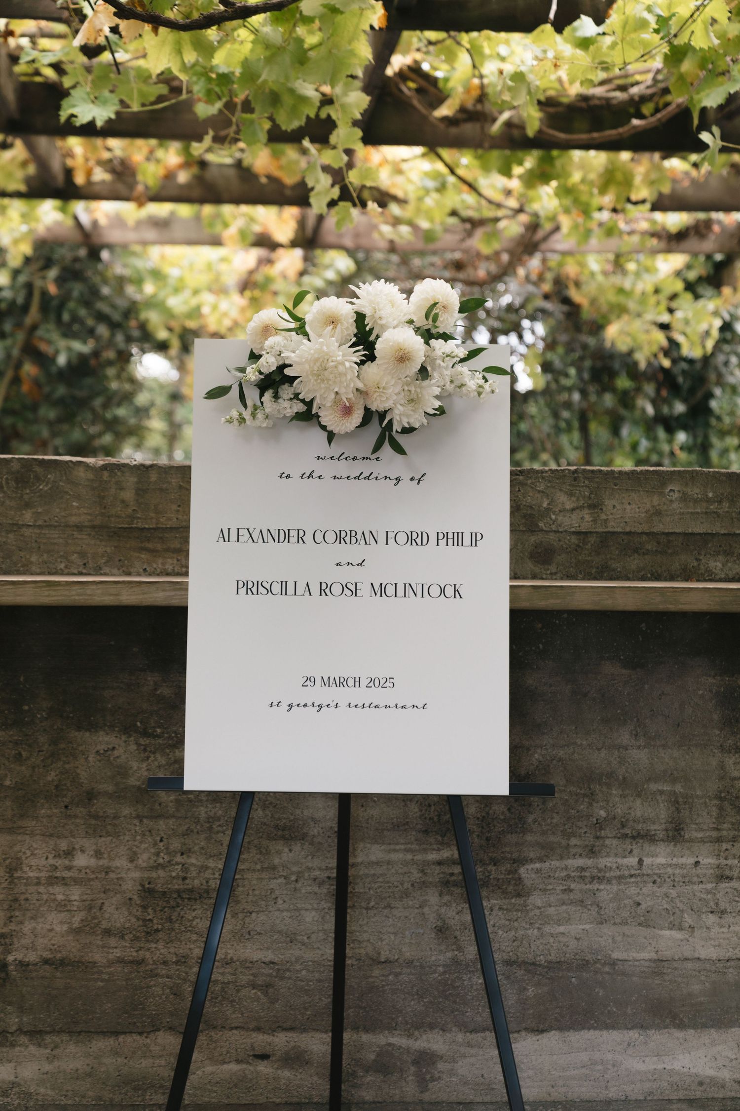 Elegant wedding welcome sign with white floral arrangements displayed on an easel in outdoor garden setting.