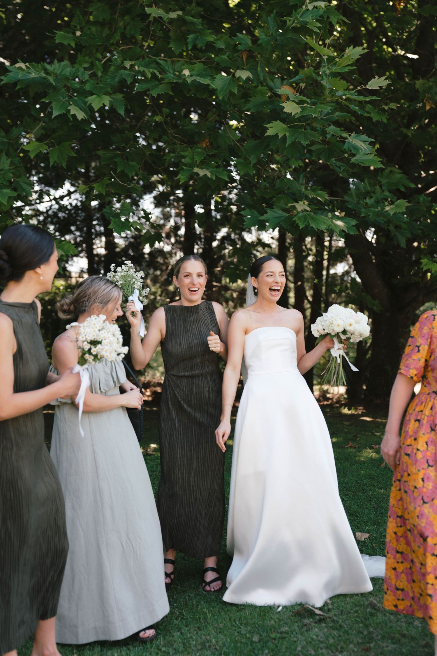 People in formal attire at an outdoor wedding celebration, with attendants in black dresses and white gowns.