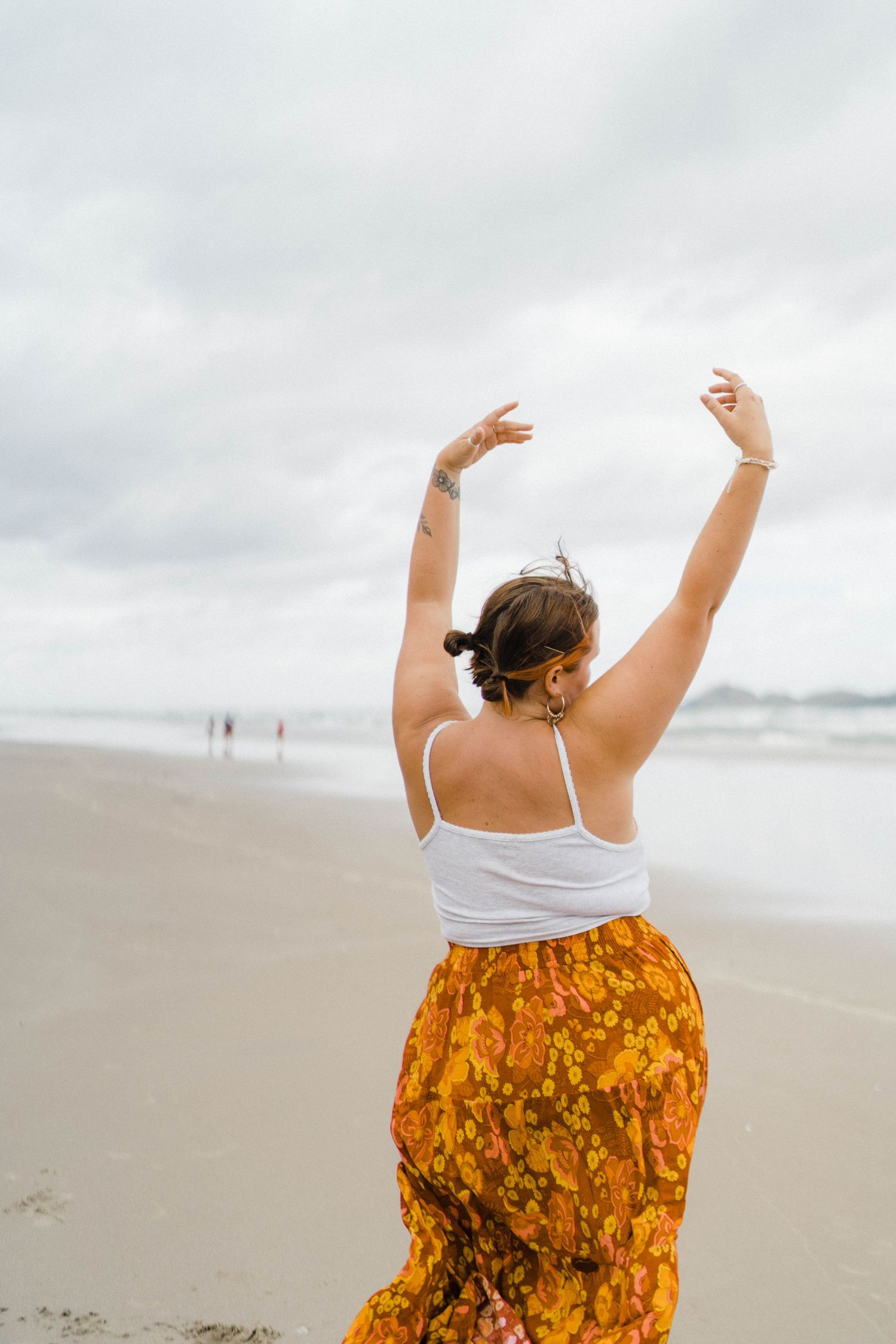 A person in a white top and flowing orange skirt dances joyfully on an overcast beach.