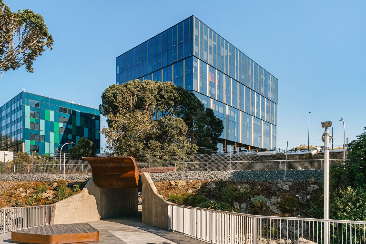 Modern glass office building with curved architectural elements and landscaping against blue sky in urban setting.