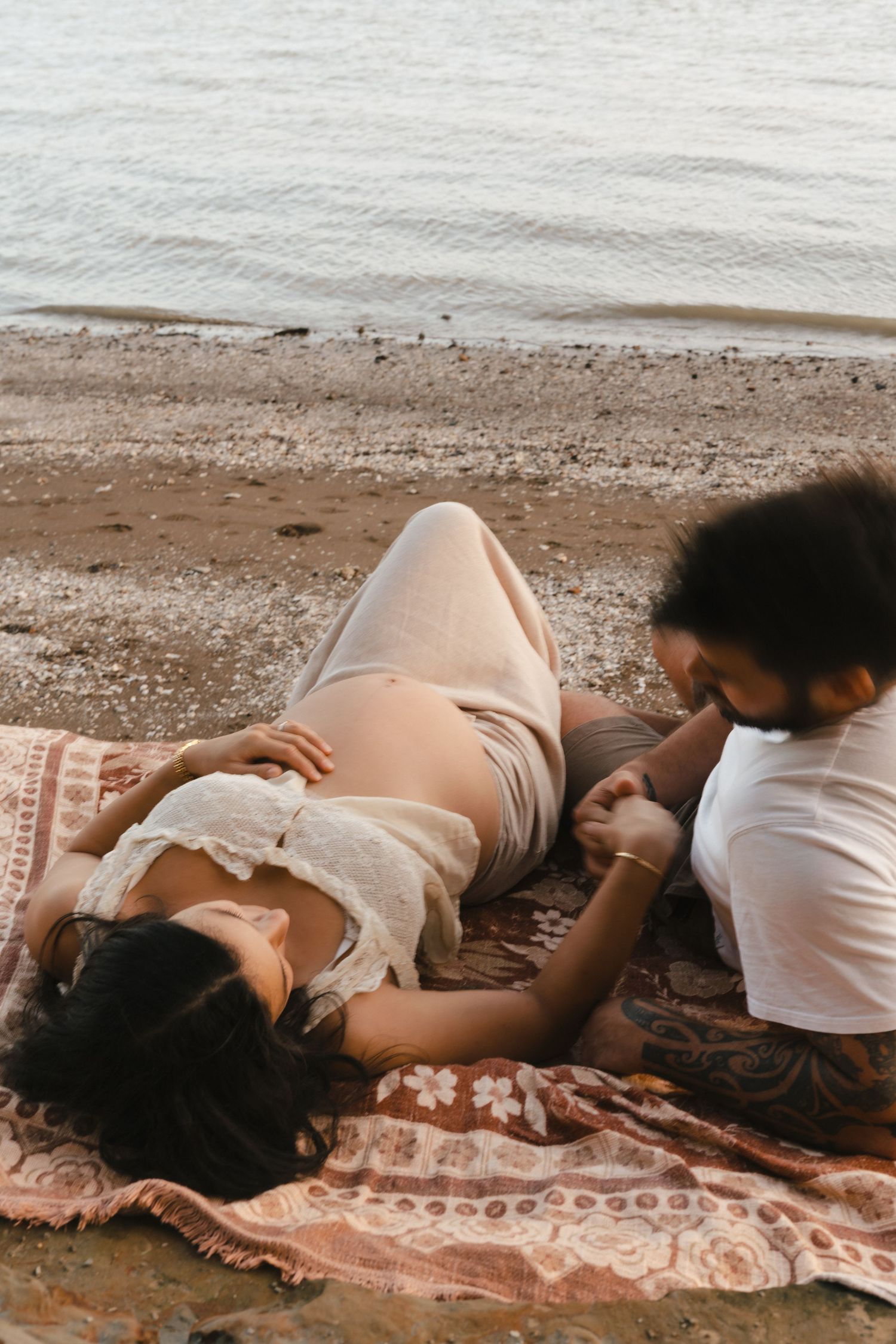 A pair relaxes together on a striped blanket at the beach while waves gently roll in near the shore.