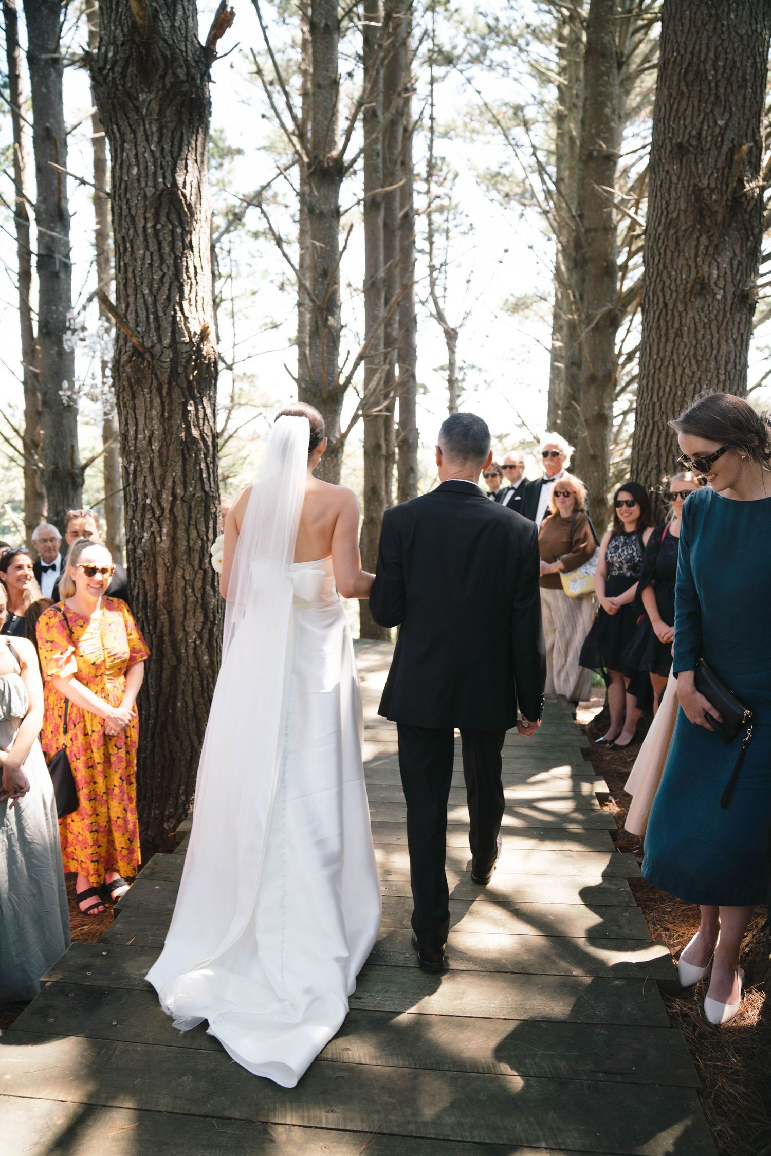 A wedding ceremony takes place on an elevated wooden pathway among tall trees.