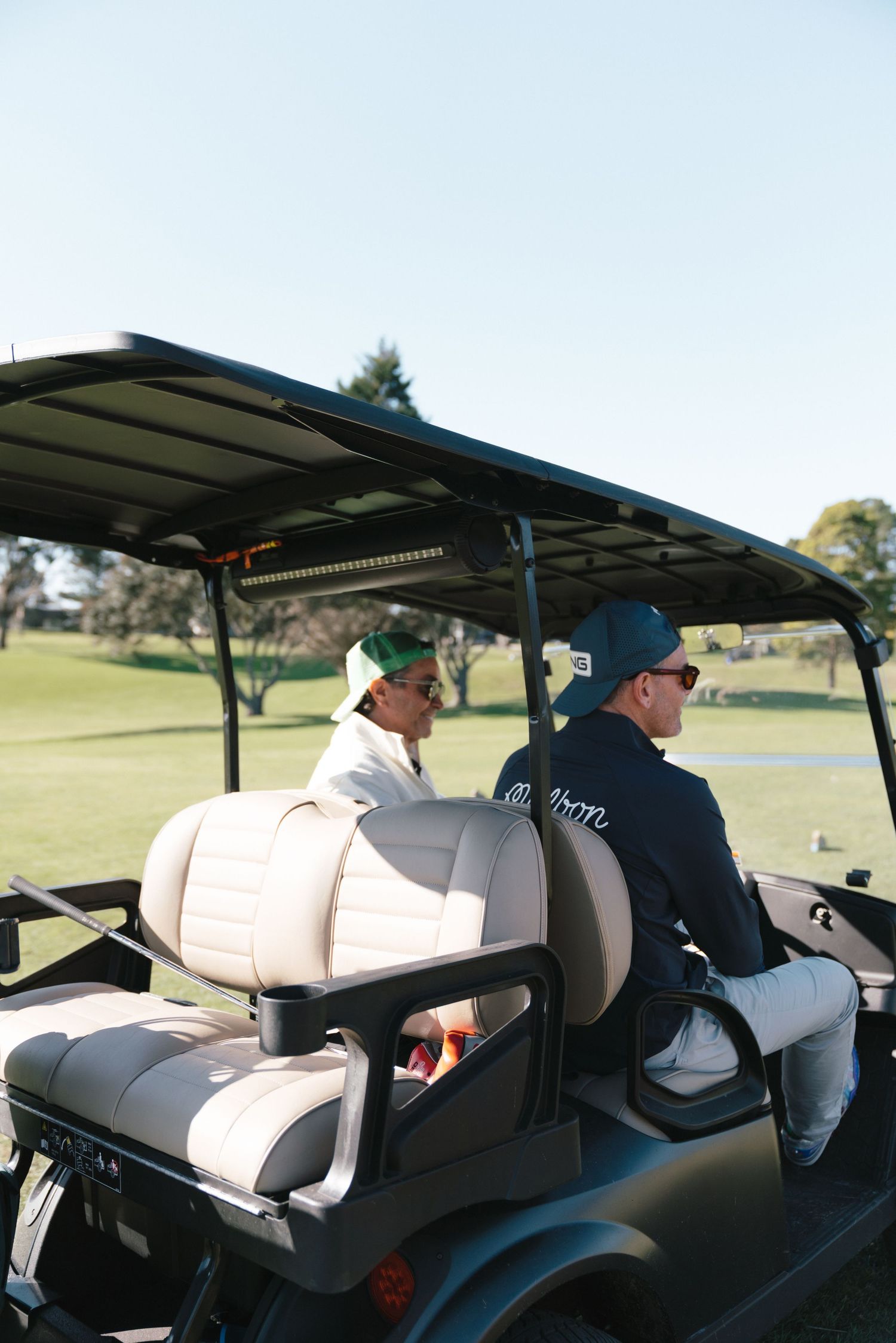 Two people riding in a golf cart on a sunny course with trees and blue sky in the background.