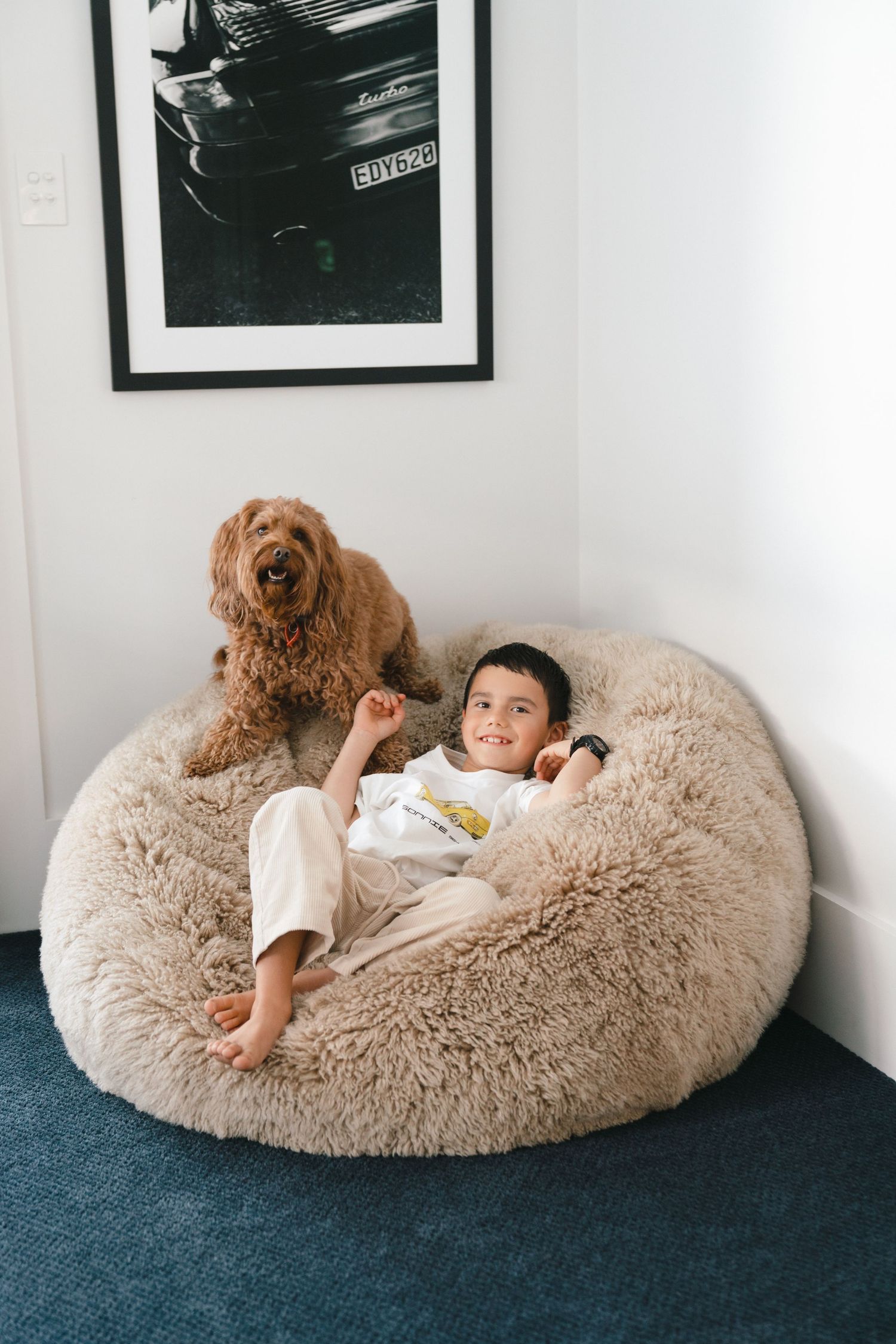 A Goldendoodle dog sits beside someone lounging on a plush beige bean bag chair in a corner with framed art.