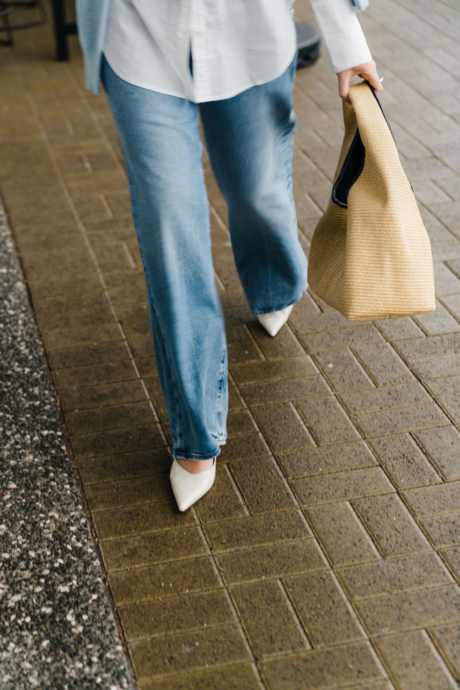 Person wearing blue jeans, white shirt and white shoes walks on brick pavement while carrying a tan woven tote bag.