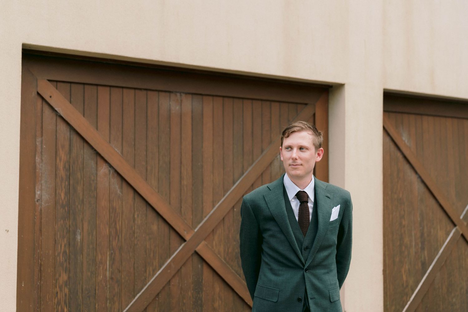 Someone in a dark green suit jacket stands in front of rustic wooden barn doors.