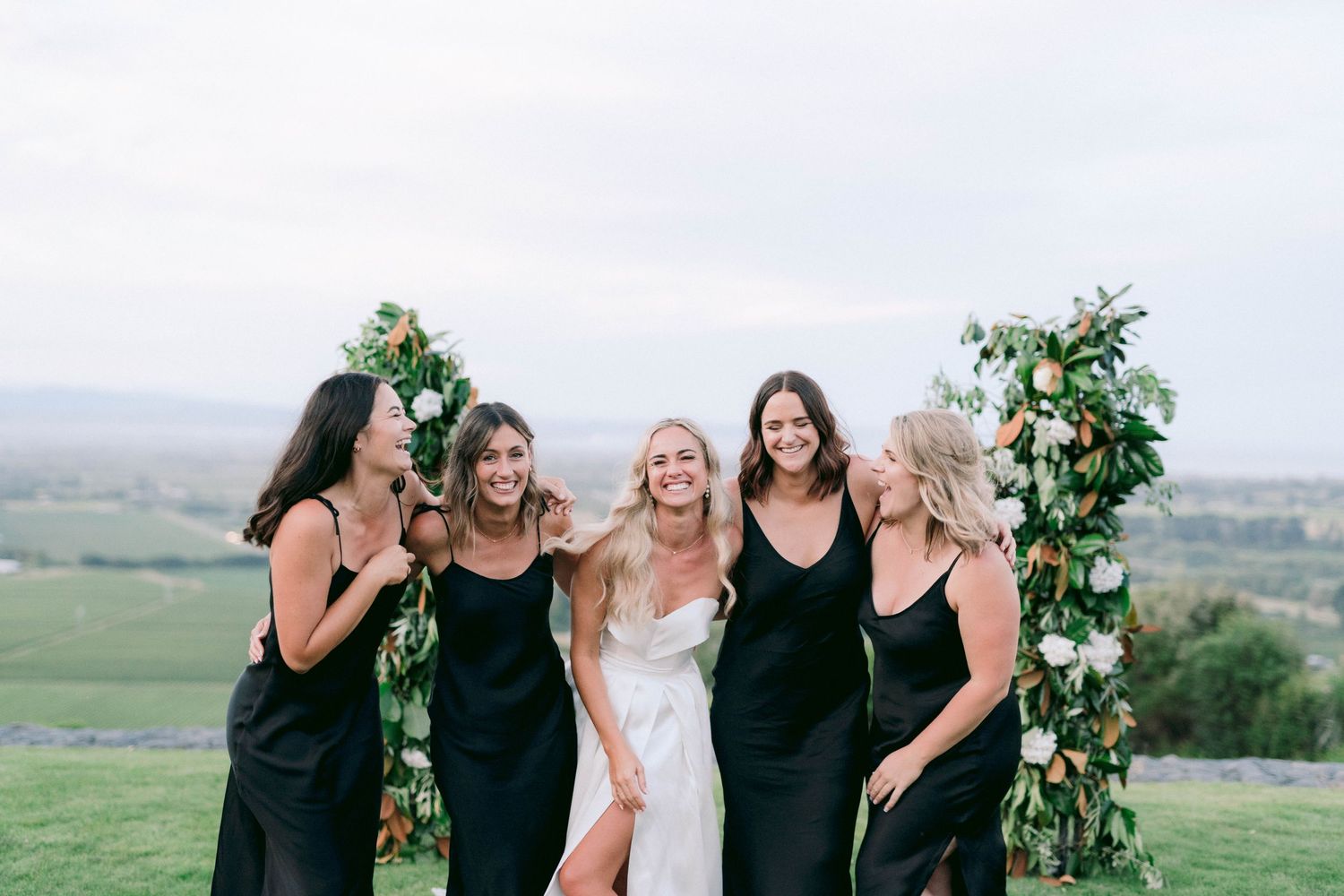 Bridal party in black dresses poses with bride against scenic countryside backdrop.