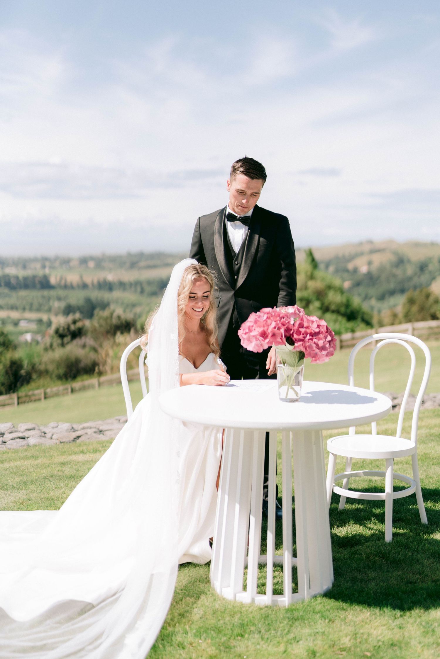 Wedding couple poses at a white outdoor table with pink roses against a scenic hilltop backdrop.