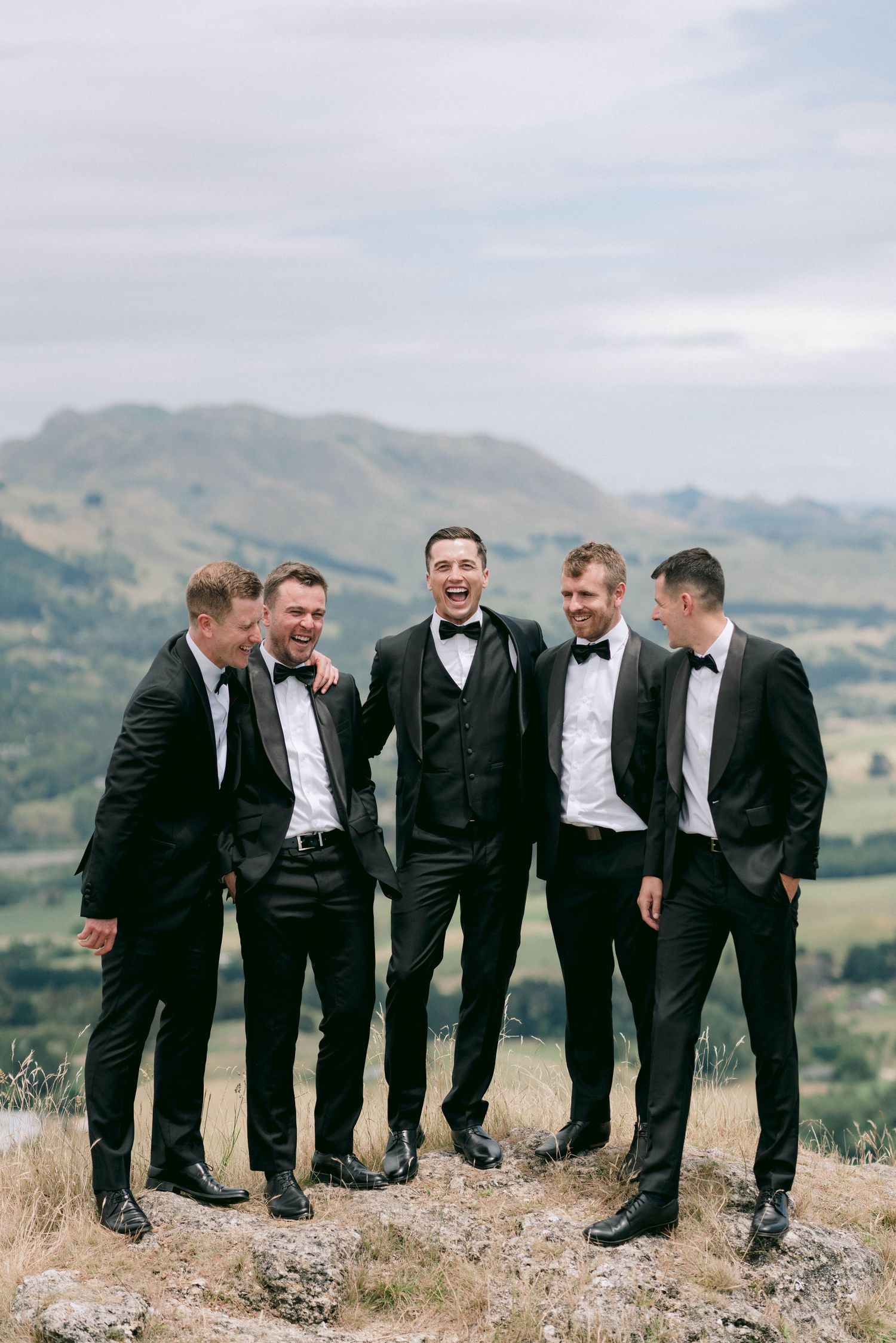 Groomsmen in black tuxedos laughing together on a hilltop with mountain views.