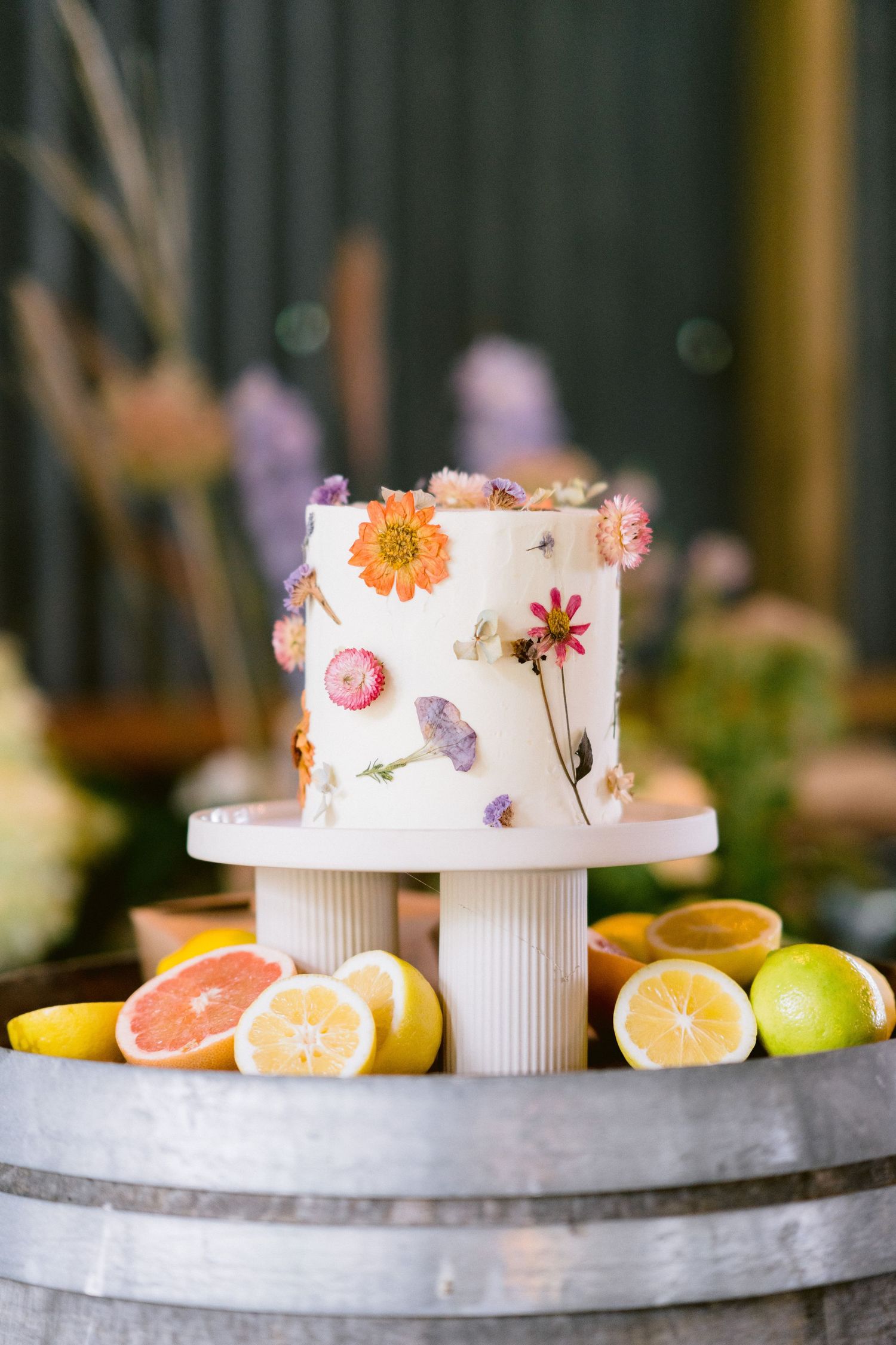 An elegant white wedding cake decorated with pressed flowers and citrus fruits displayed on a concrete counter.