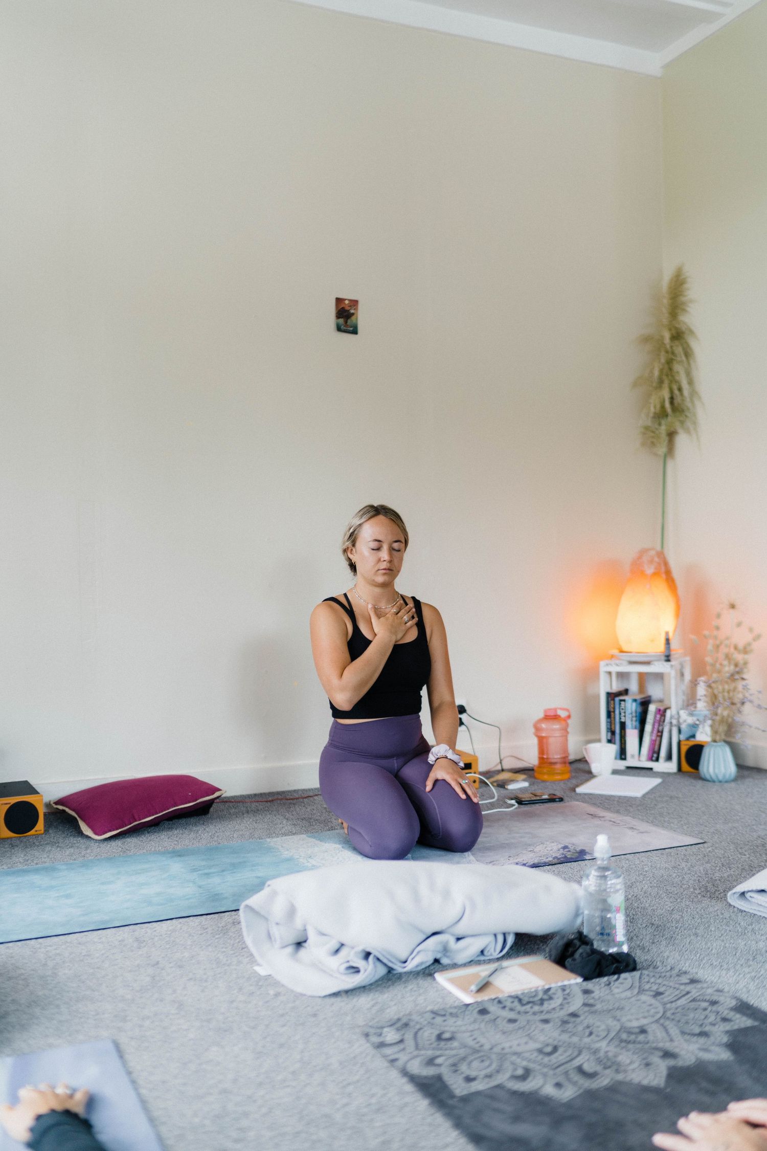 A yoga practitioner in purple leggings sits on a mat with candles and wellness items nearby.