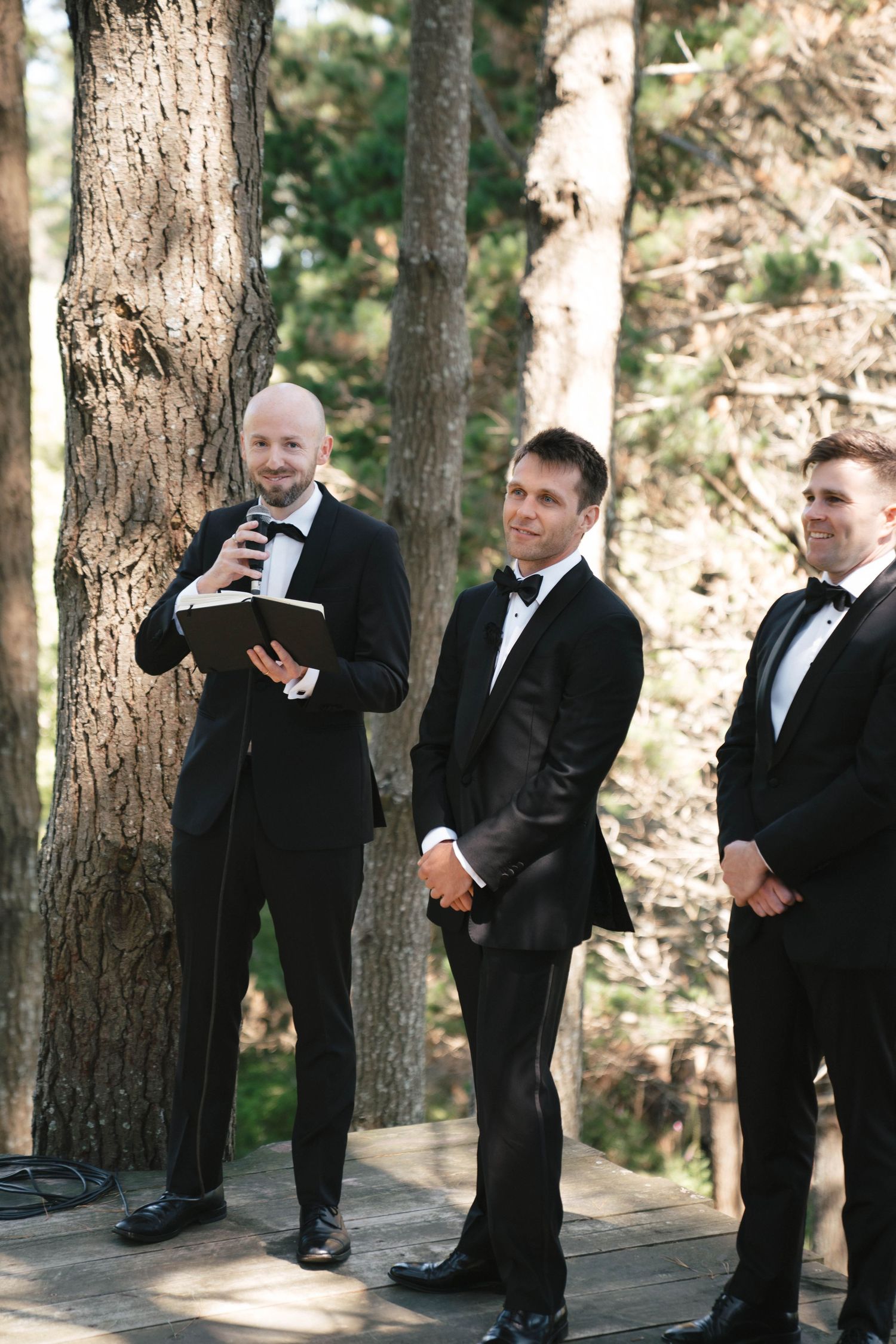 Three men in formal tuxedos stand on a wooden platform during an outdoor ceremony.