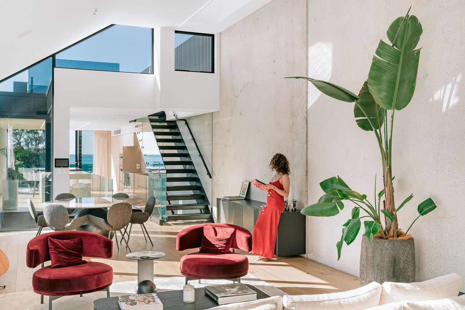 Contemporary white interior showcases a red accent chair and tropical plant beside a glass staircase.