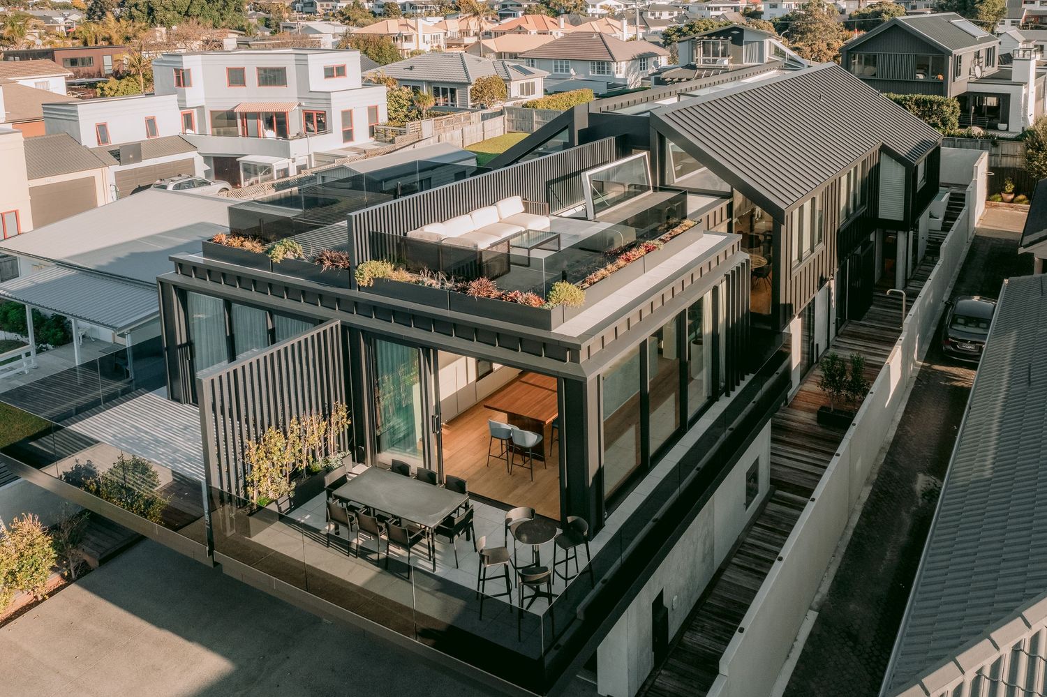 Modern rooftop terrace with glass railings and outdoor seating overlooking a residential neighborhood at sunset.