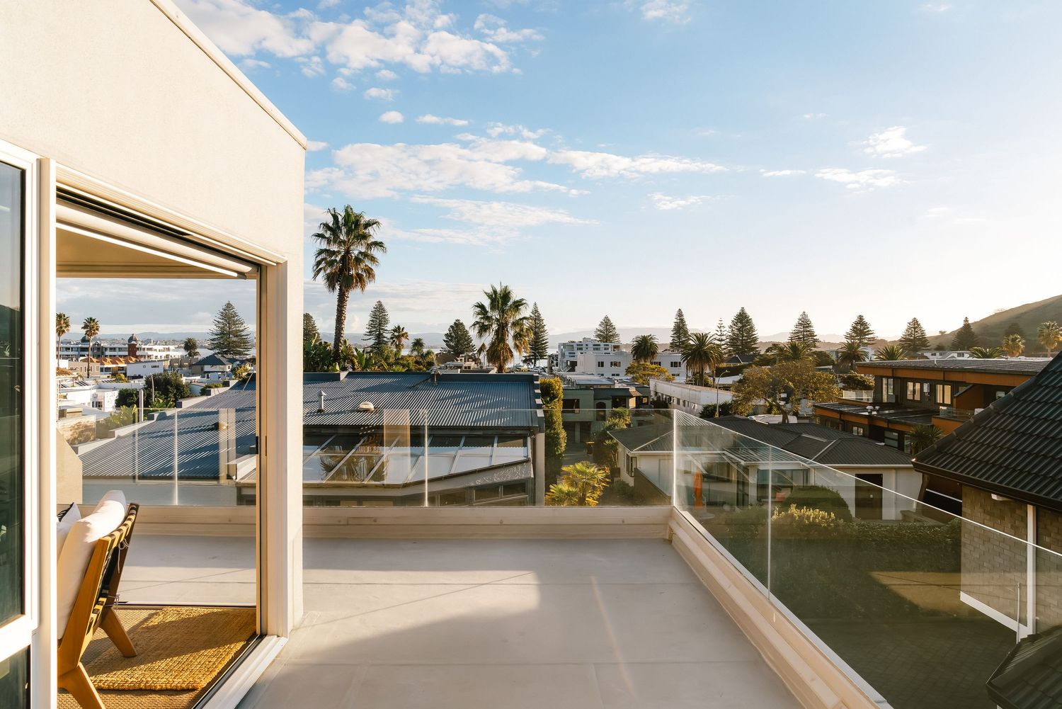 Panoramic rooftop terrace with glass railings overlooks coastal neighborhood under blue skies.