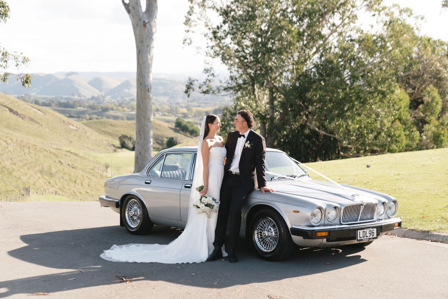 Newlywed couple posing with classic silver Jaguar car in scenic countryside.