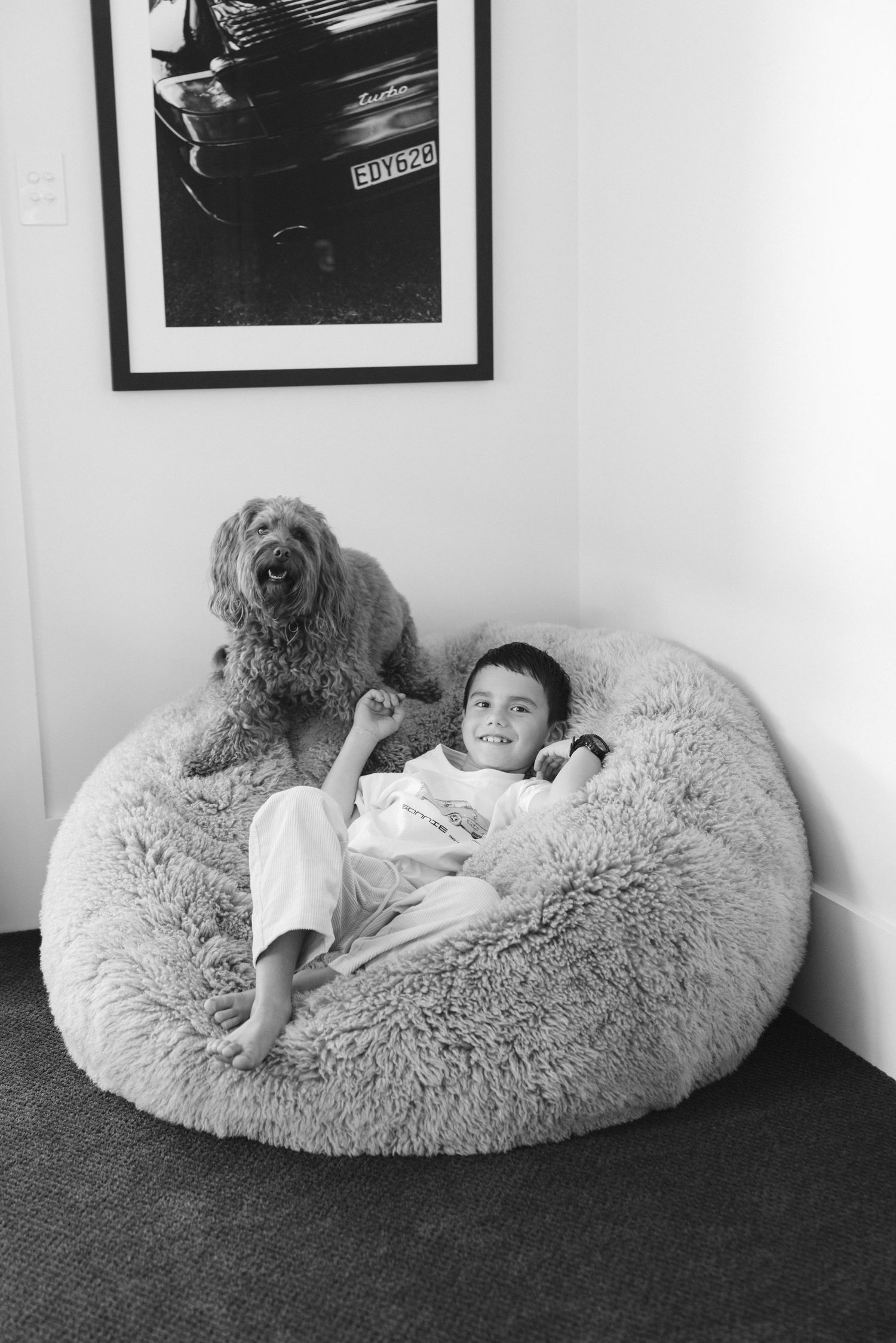 A Goldendoodle dog lounges next to a child on a plush round bean bag chair in a black and white photograph.
