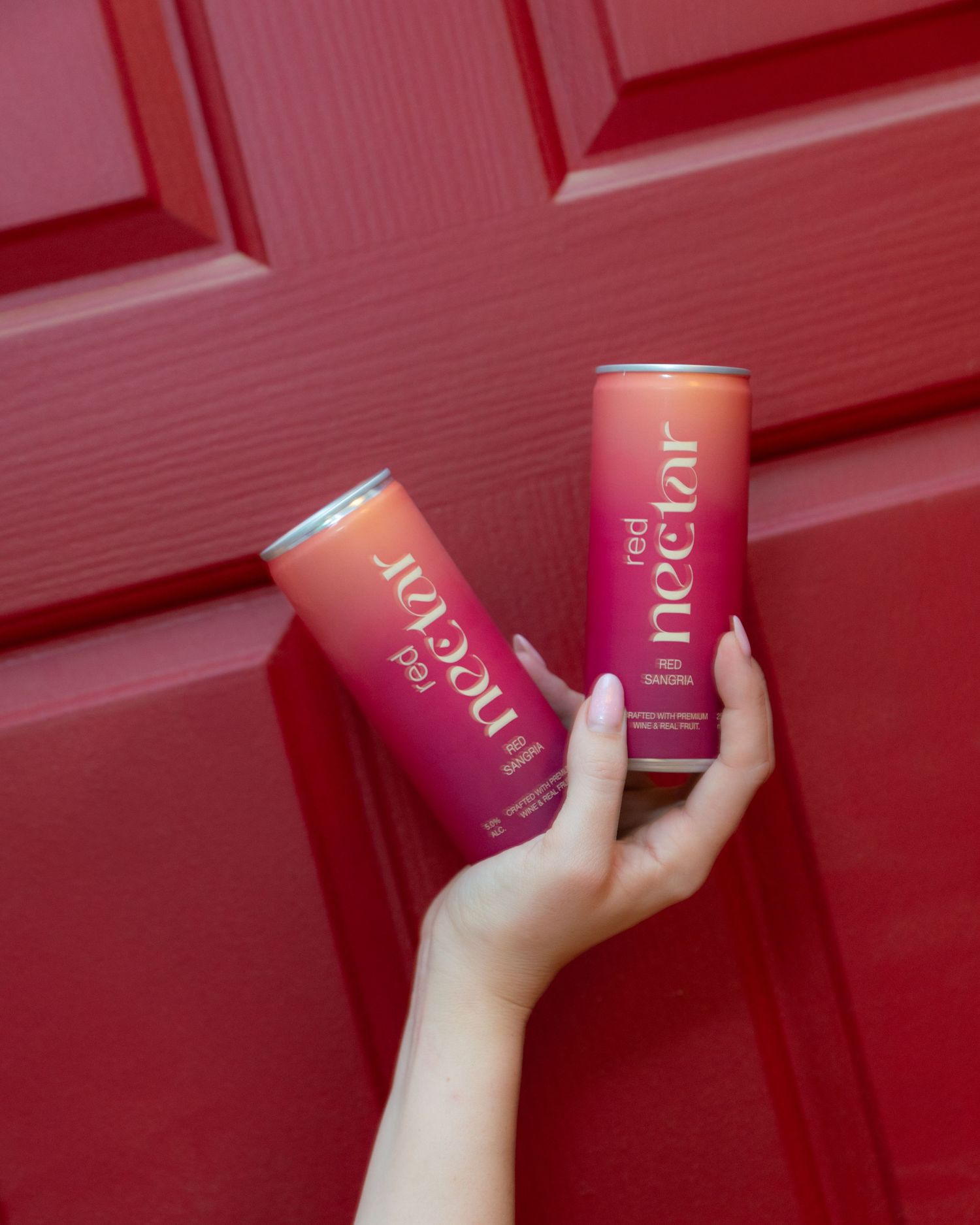 Hand holding two pink cans of Poppi prebiotic soda against a red door background.