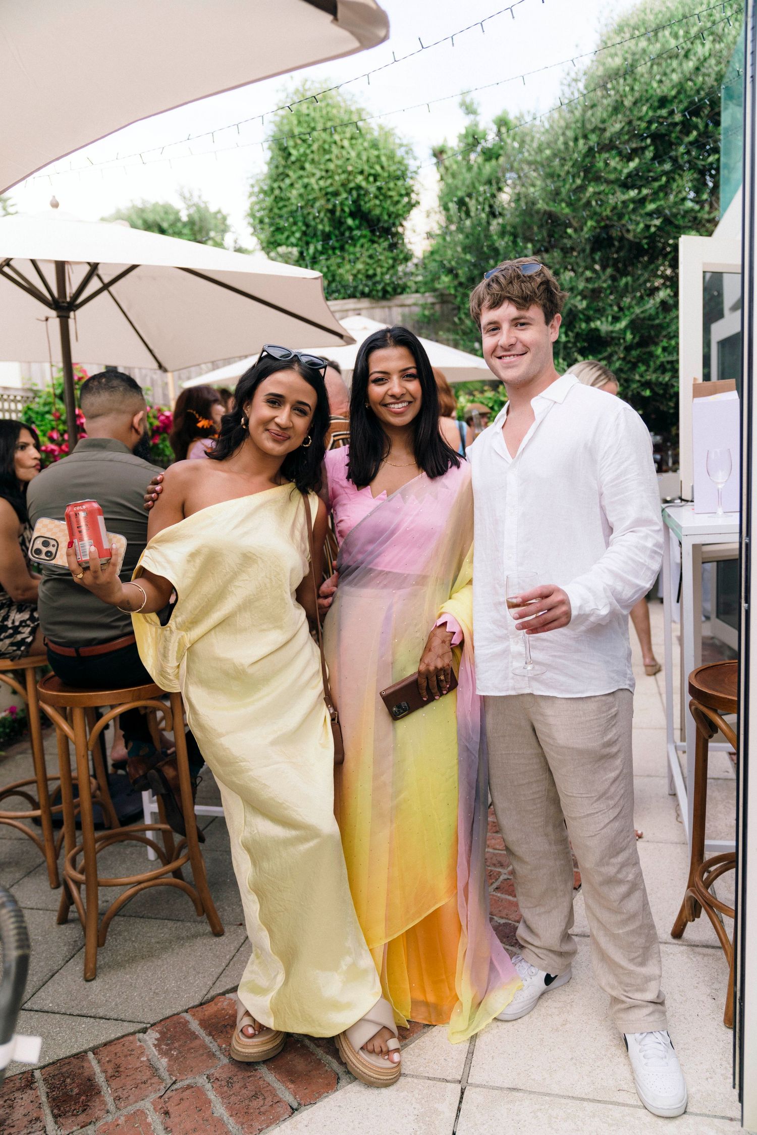A group of people in light summer attire celebrate at an outdoor party venue with umbrellas and seating.