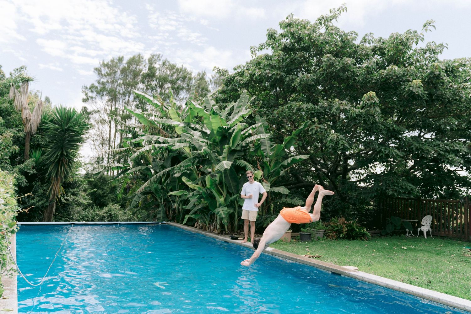 Swimming pool with diving board surrounded by lush greenery and multiple people enjoying the water.
