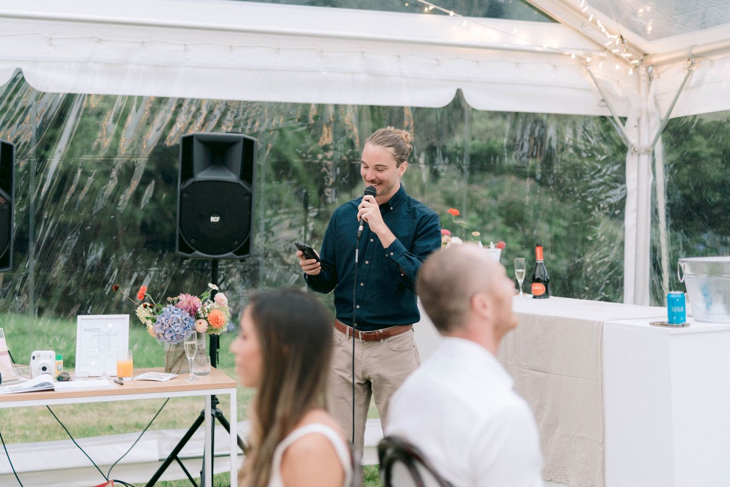 A speaker gives a speech under a white tent during an outdoor celebration with guests seated at tables.