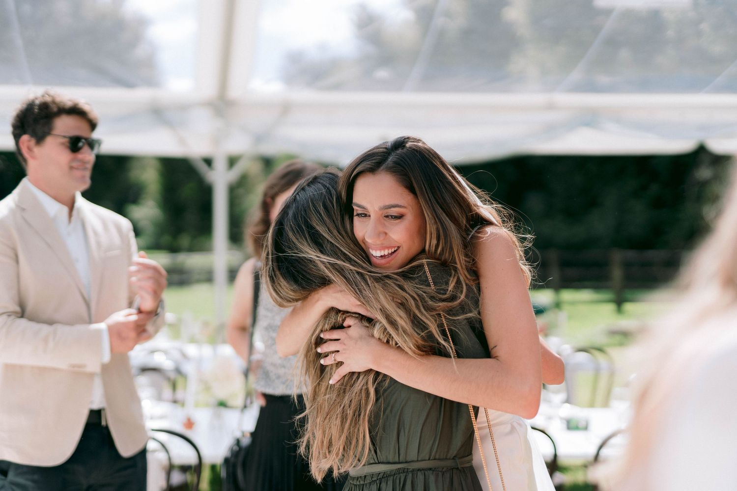Wedding attendees embrace and celebrate during an outdoor reception under white tents.