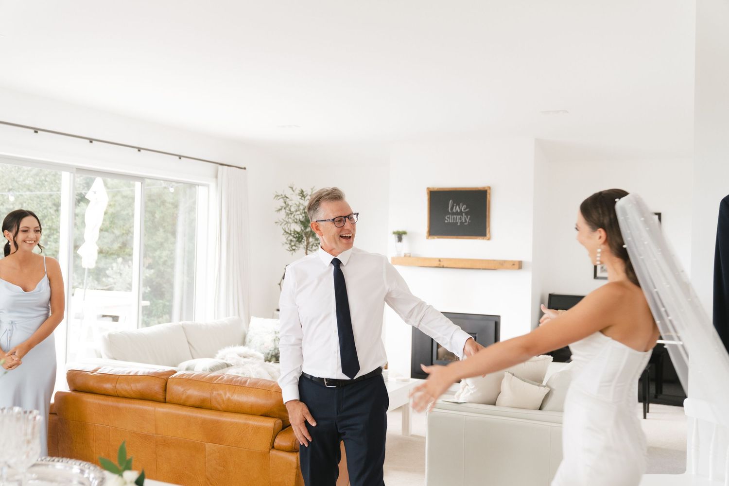 A family in formal attire greets each other in a bright white living room with leather sofa and piano.
