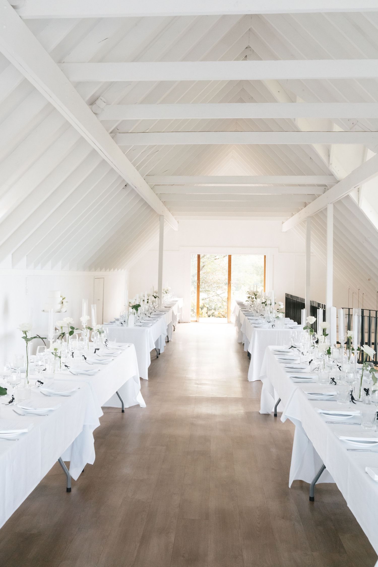 White reception hall decorated with elegant table settings and crisp linens under exposed wooden beams and natural lighting.