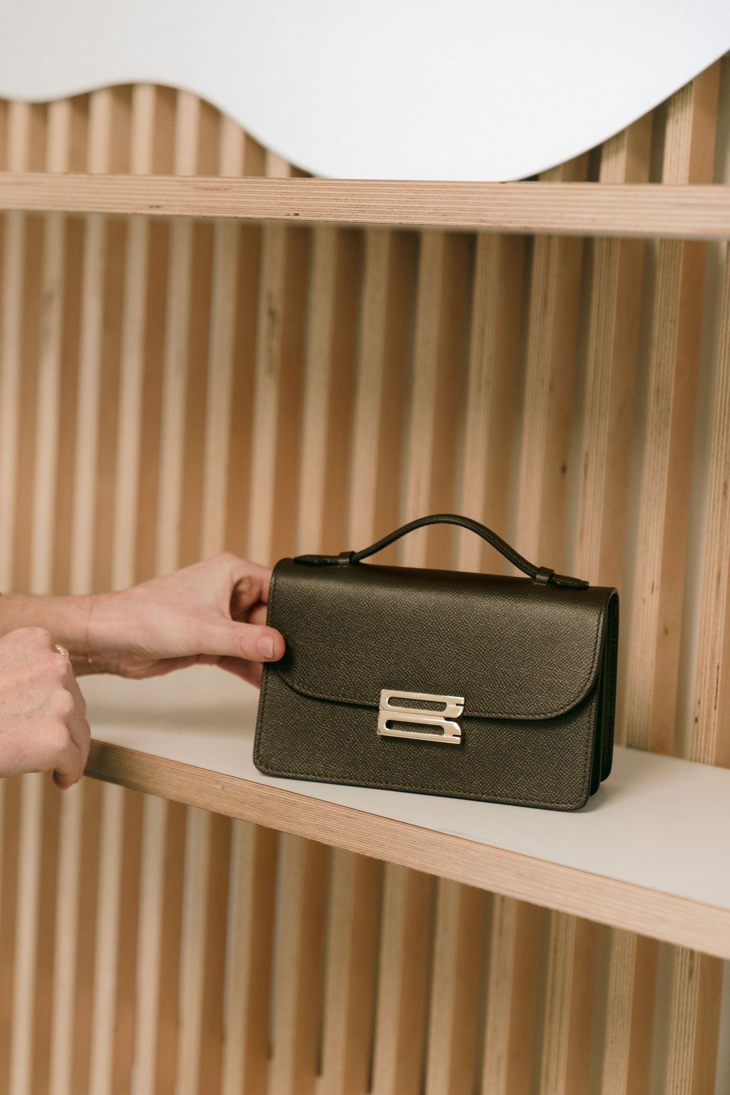 Hand reaches for a sleek black leather handbag with gold hardware displayed on a wooden shelf with ribbed wall background.