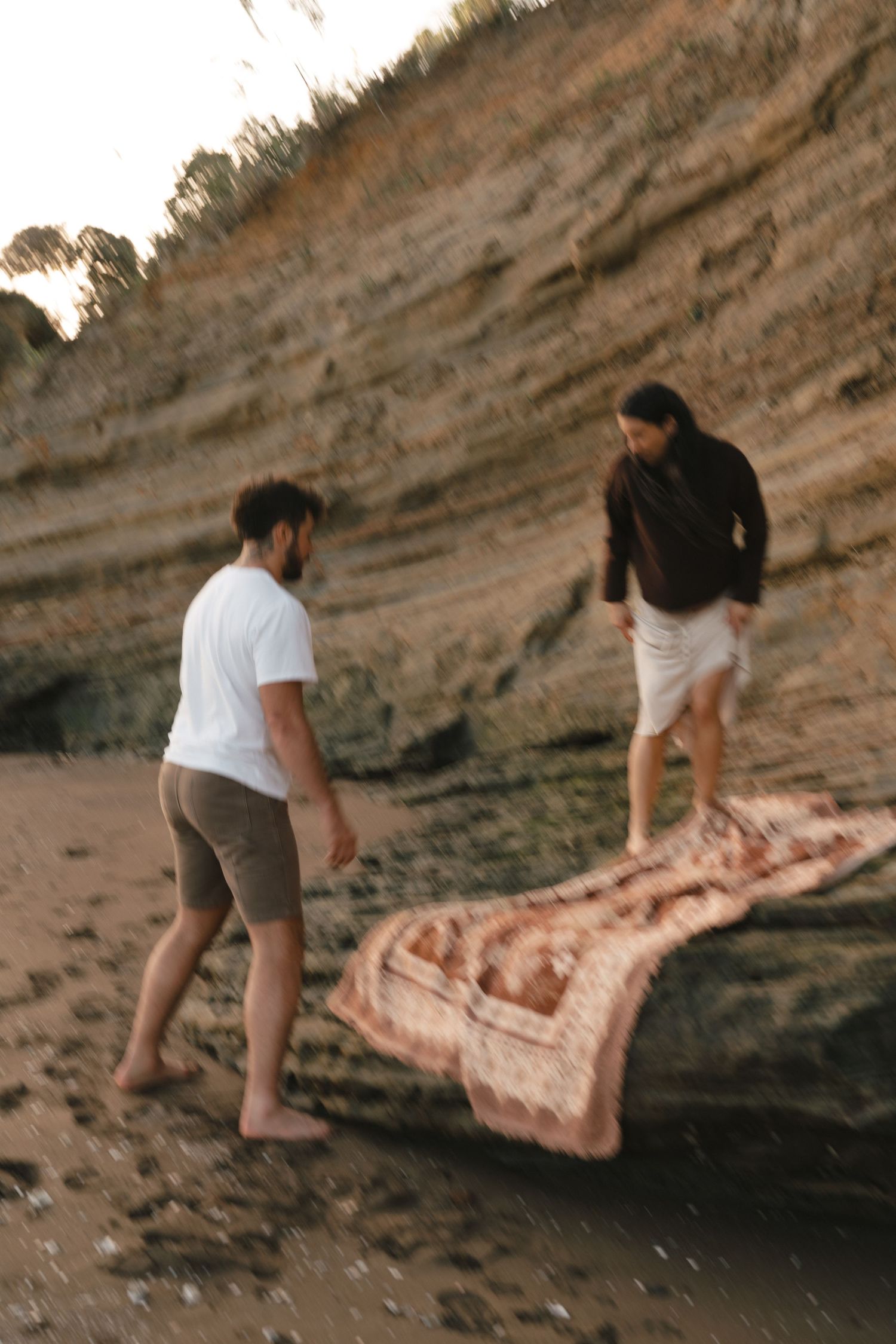 Two people setting up blanket for picnic near rocky coastal cliffs at sunset.