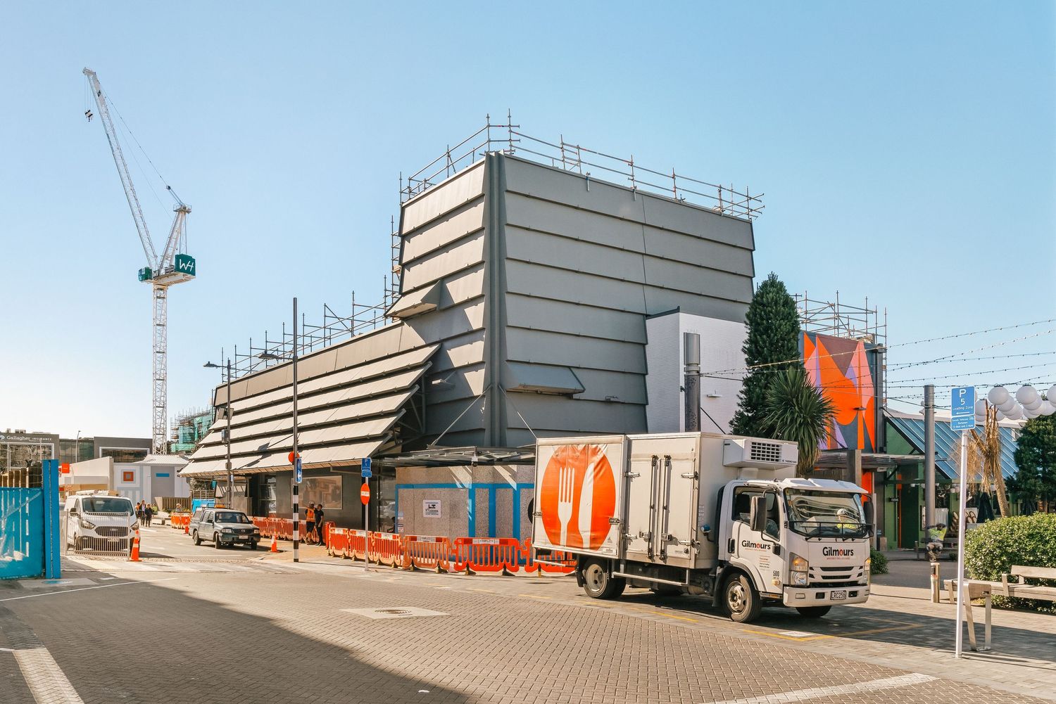 Construction site shows multiple industrial buildings and equipment with orange barriers and safety railings visible.