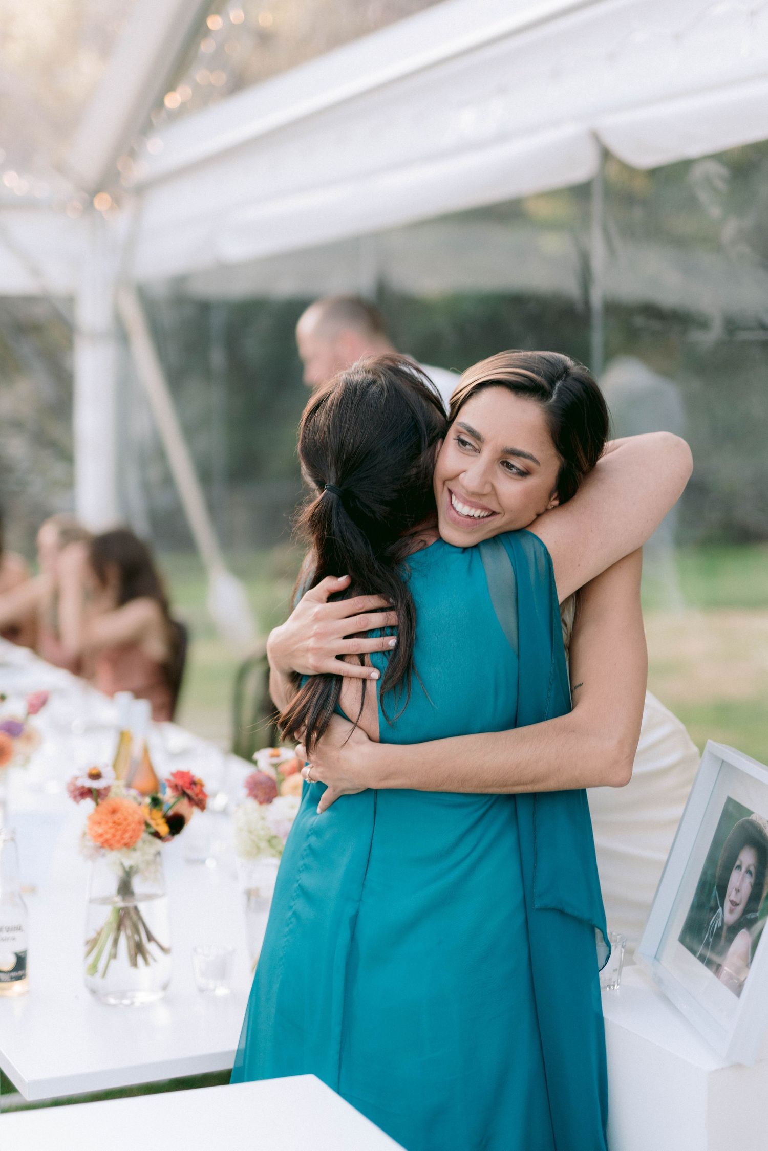 Two women share an emotional embrace at a wedding reception under a white tent.