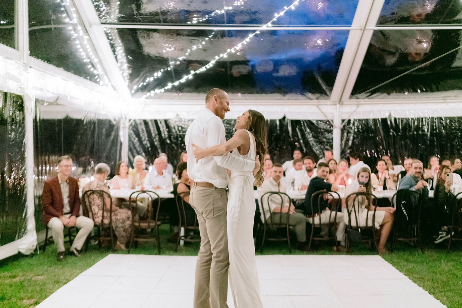 String lights illuminate an outdoor wedding reception under a tent at dusk.