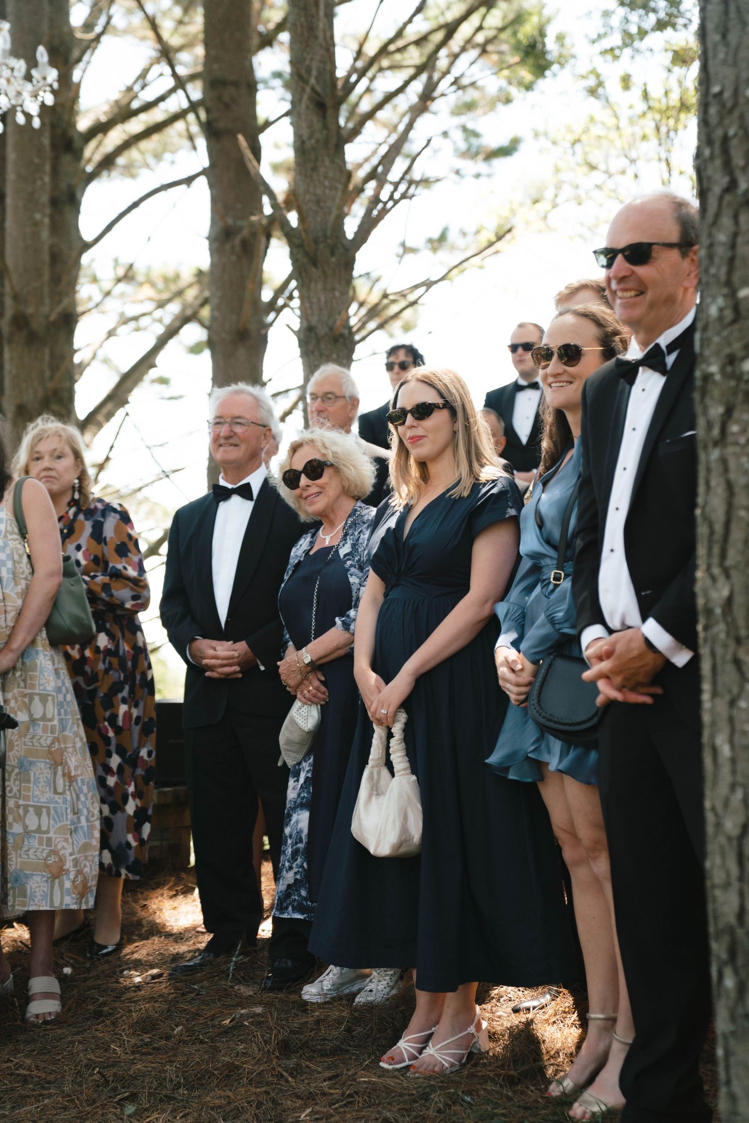 Wedding guests in formal attire gathered outdoors among pine trees.