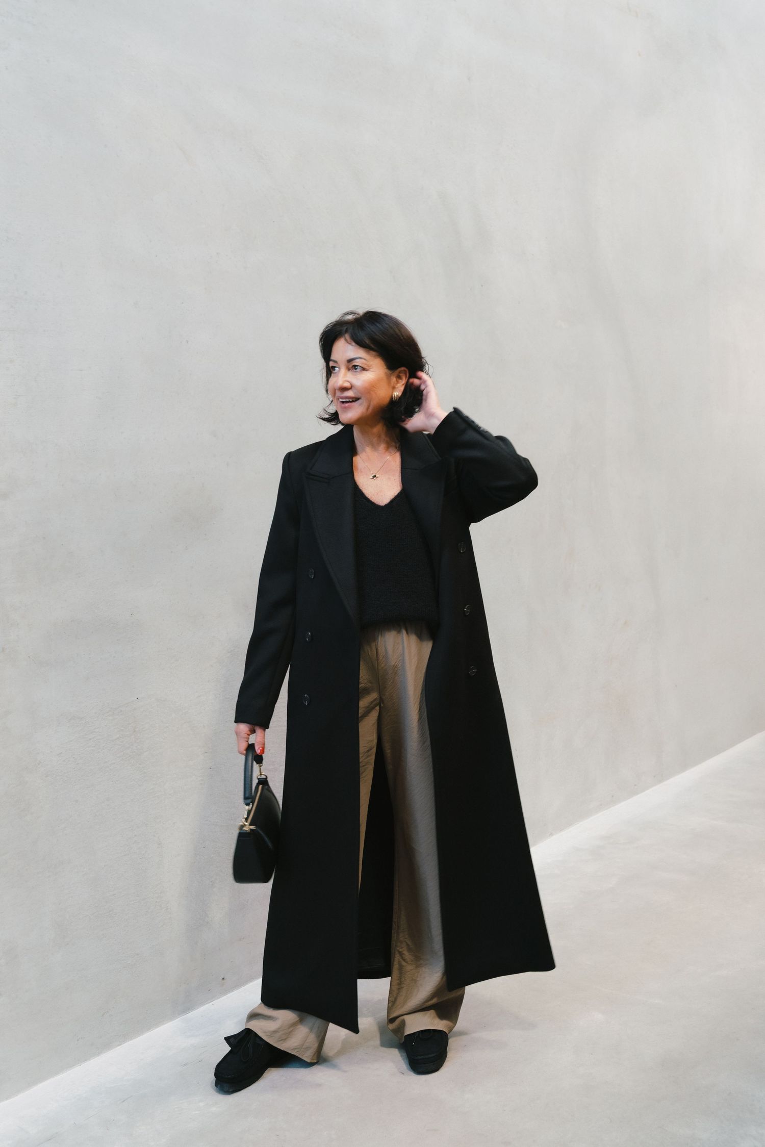 Elegant minimalist outfit featuring long black coat, beige trousers and black handbag against white wall.