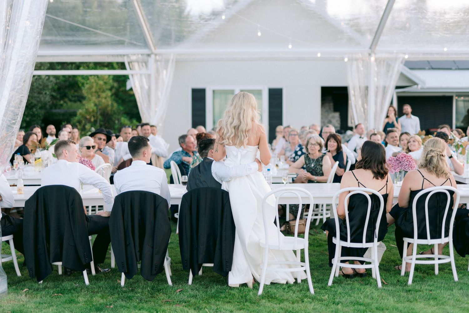 Wedding reception setup outdoors with white chairs and tables under tent lighting.