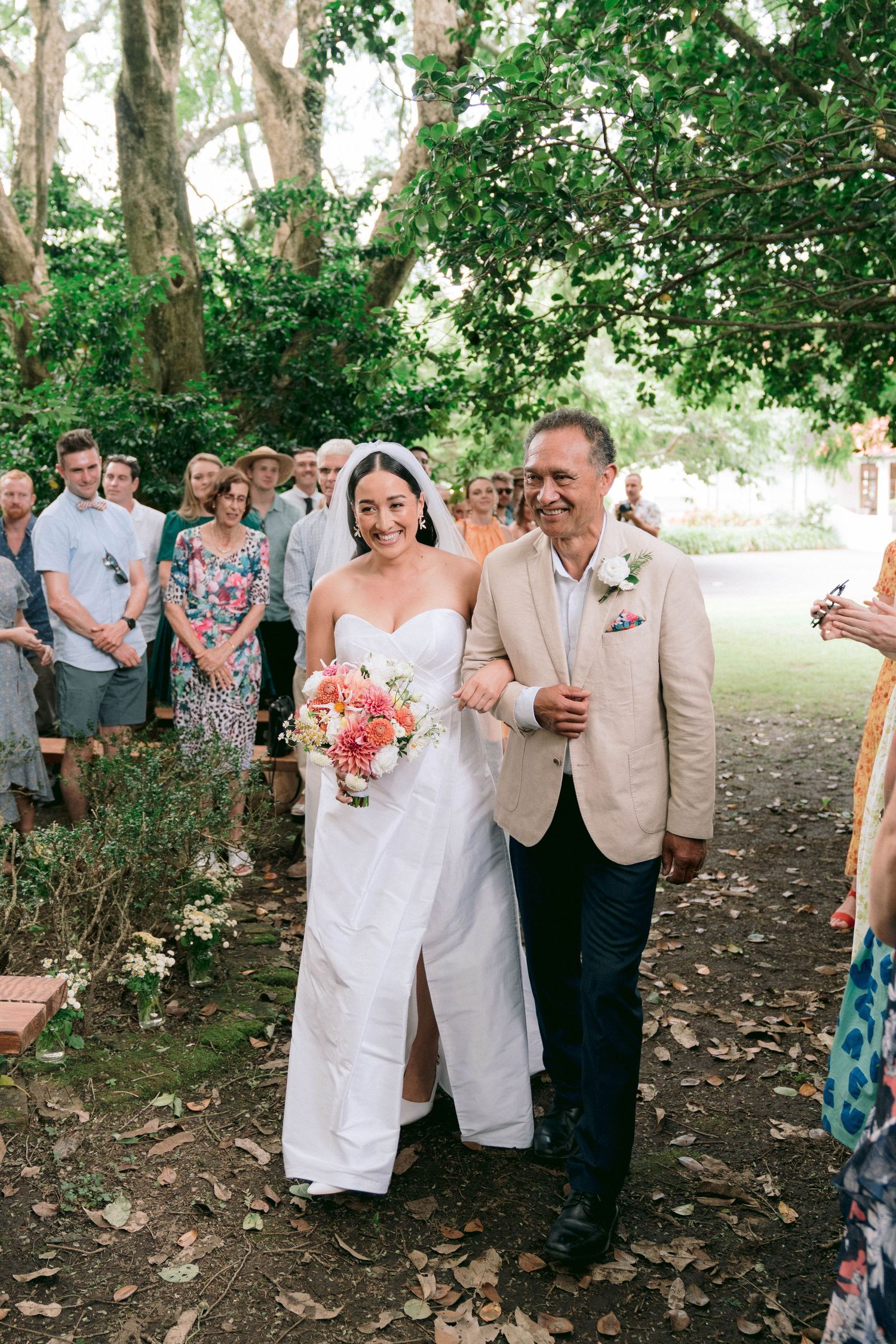 Wedding ceremony processional under tree canopy with guests watching the couple walk down leaf-covered aisle.