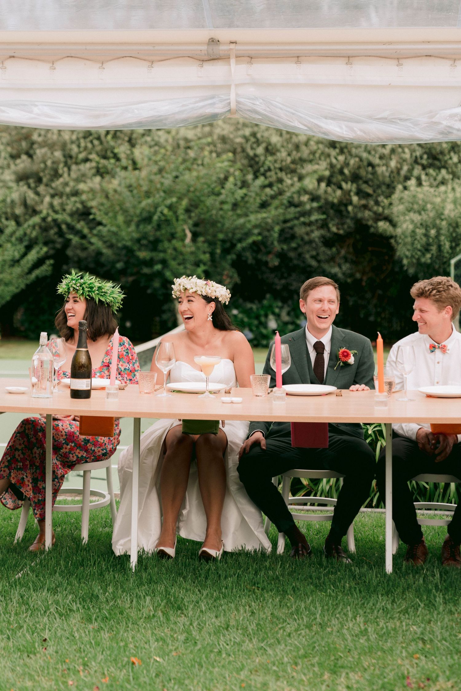 Wedding party members sit at a banquet table outdoors under a white tent during a summer celebration.