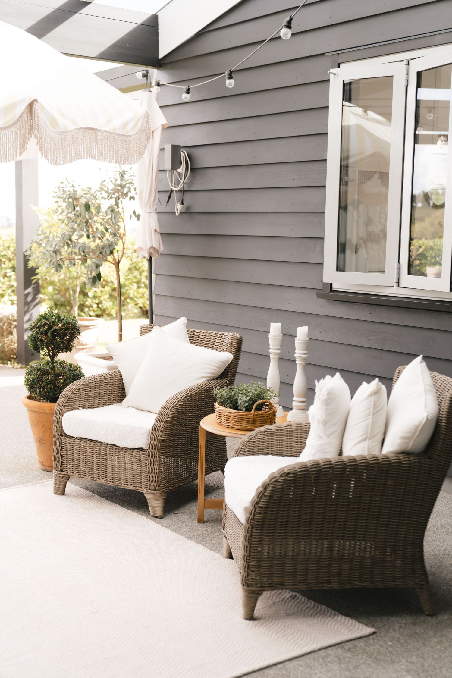Cozy outdoor patio seating area with wicker chairs, white cushions and pillows against grey house siding.