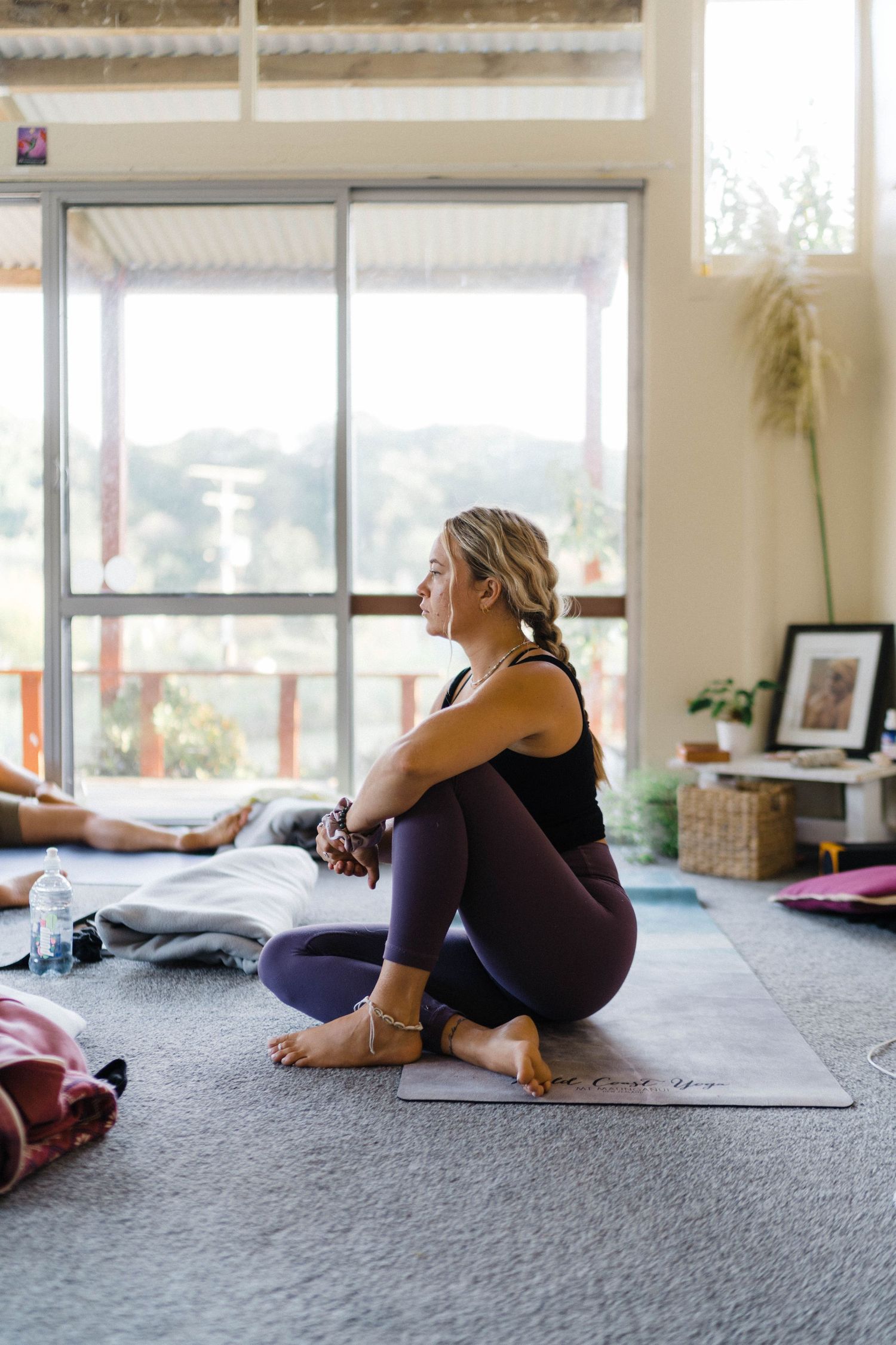 Yoga practitioner in purple leggings sits in a relaxed pose on a mat by large windows.