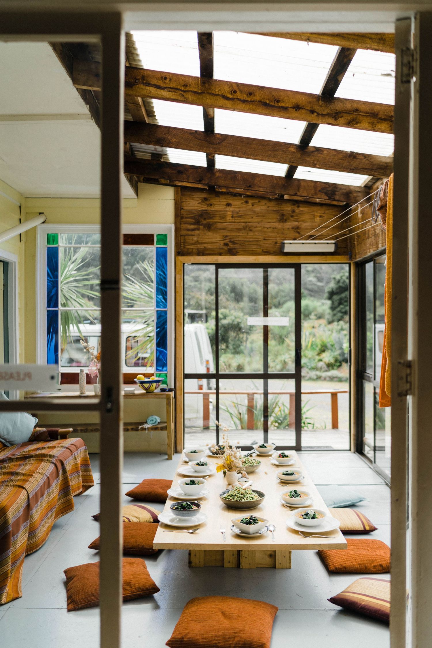 Modern dining room with wooden ceiling beams, large windows, and orange cushioned bench seating around a long table.