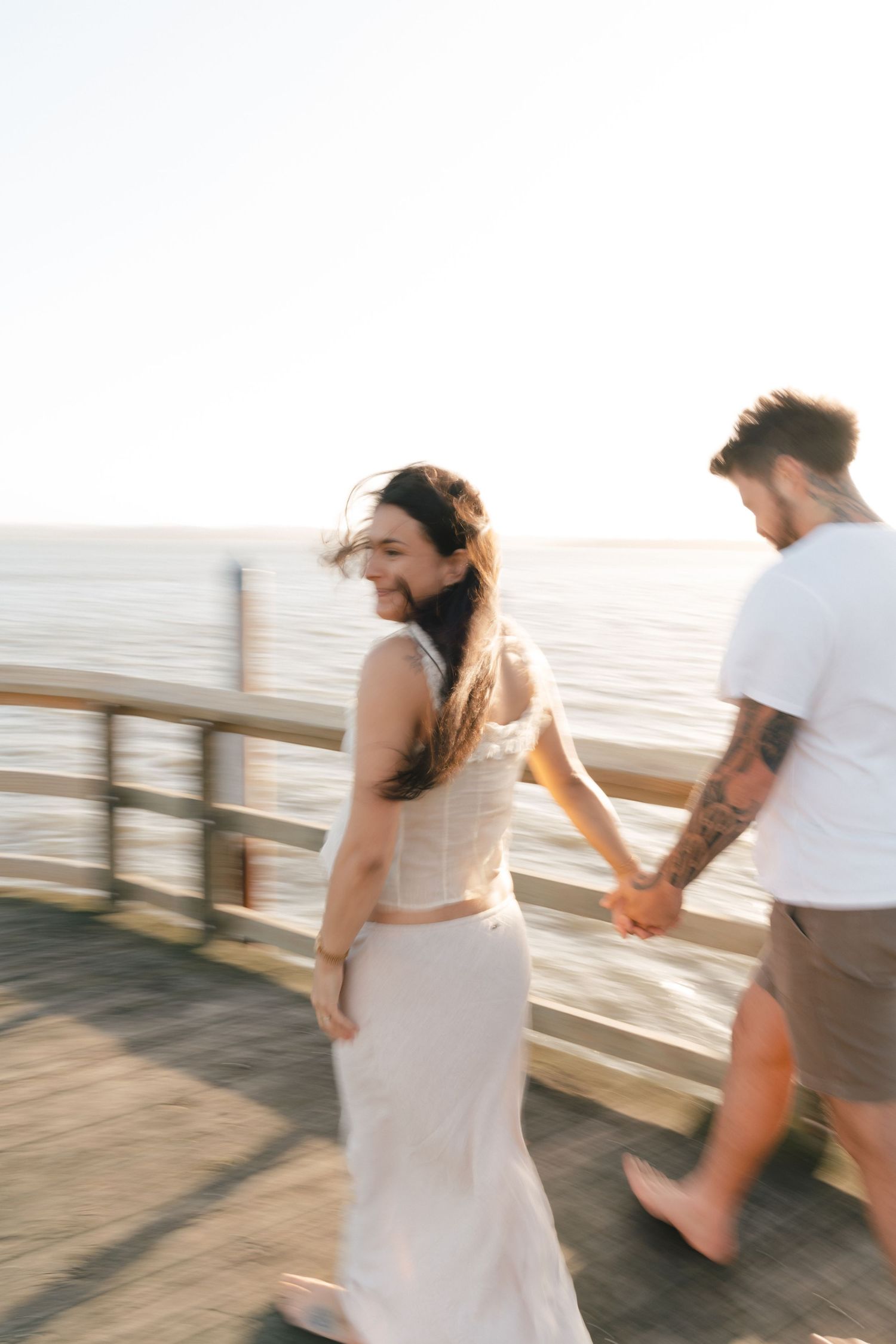 Couple holding hands while walking on a wooden pier at sunset by the ocean in casual summer attire.