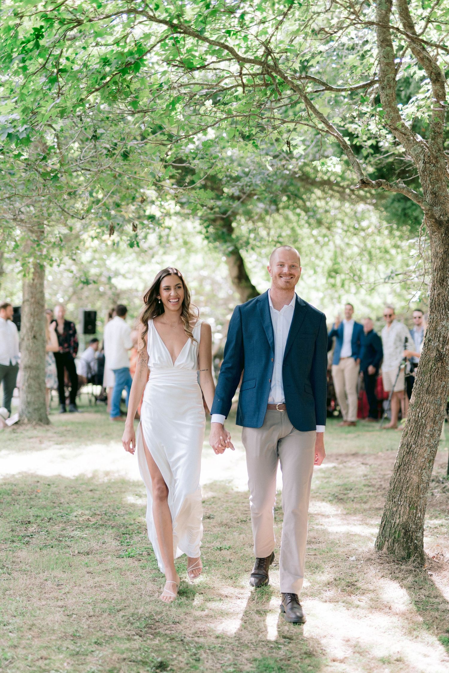 A couple walks hand in hand through a tree-lined path at an outdoor wedding celebration.
