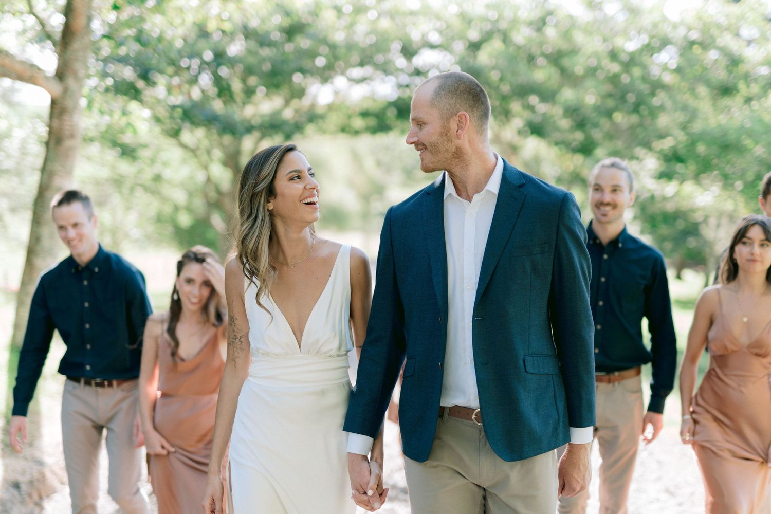 Wedding party members walking and laughing together on a tree-lined path.